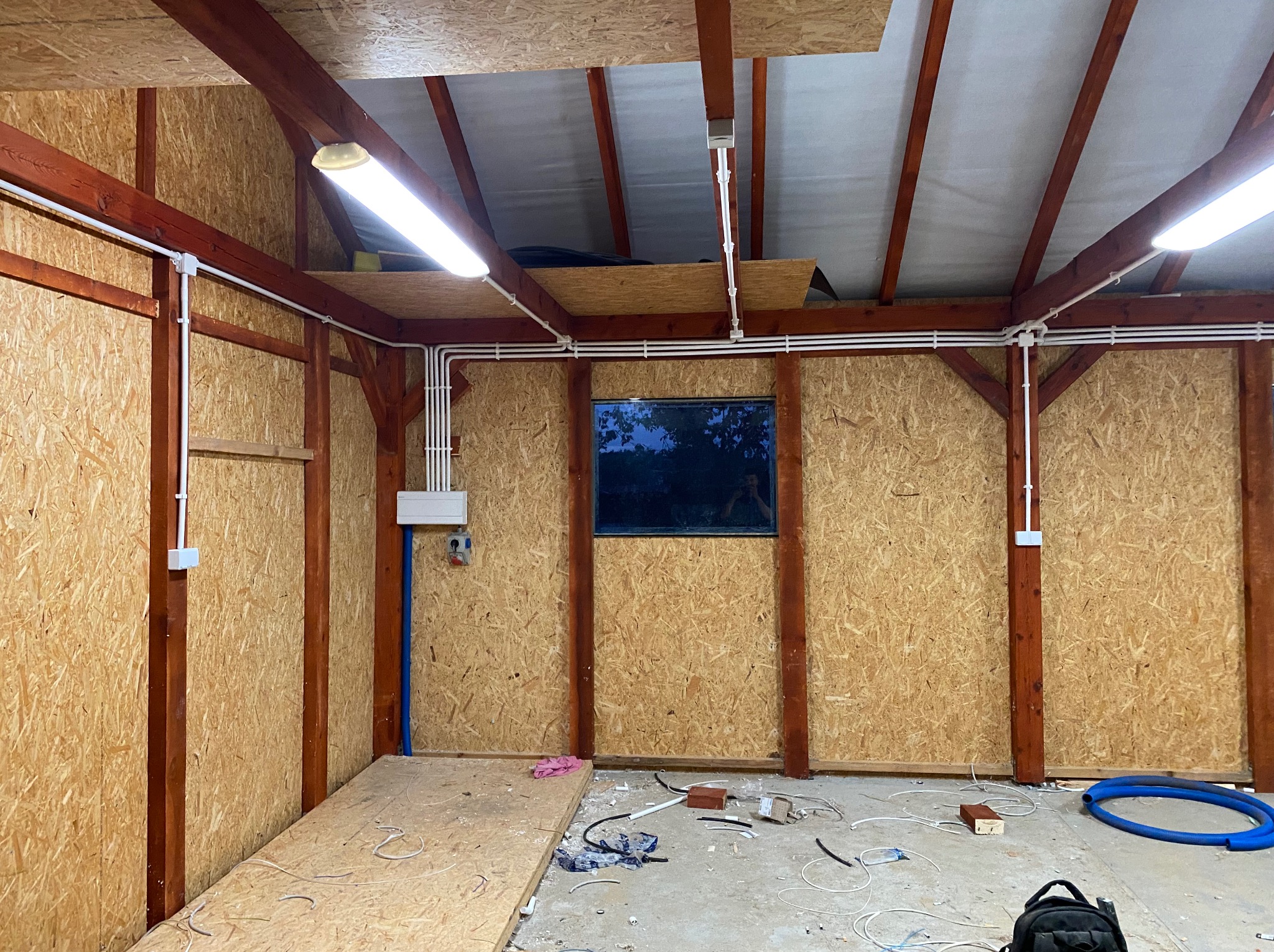 Wnętrze garażu lub warsztatu w trakcie prac elektrycznych: widoczne kable w białych rurkach, skrzynka elektryczna, drewniana konstrukcja ścian i sufitu z płyt OSB, okno w tle, oraz porozrzucane...