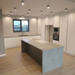 Nowoczesna kuchnia z wyspą: białe szafki, szary blat, płyta indukcyjna. Minimalistyczny design, oświetlenie punktowe, okno z widokiem na zewnątrz. Betonowa wyspa kuchenna.