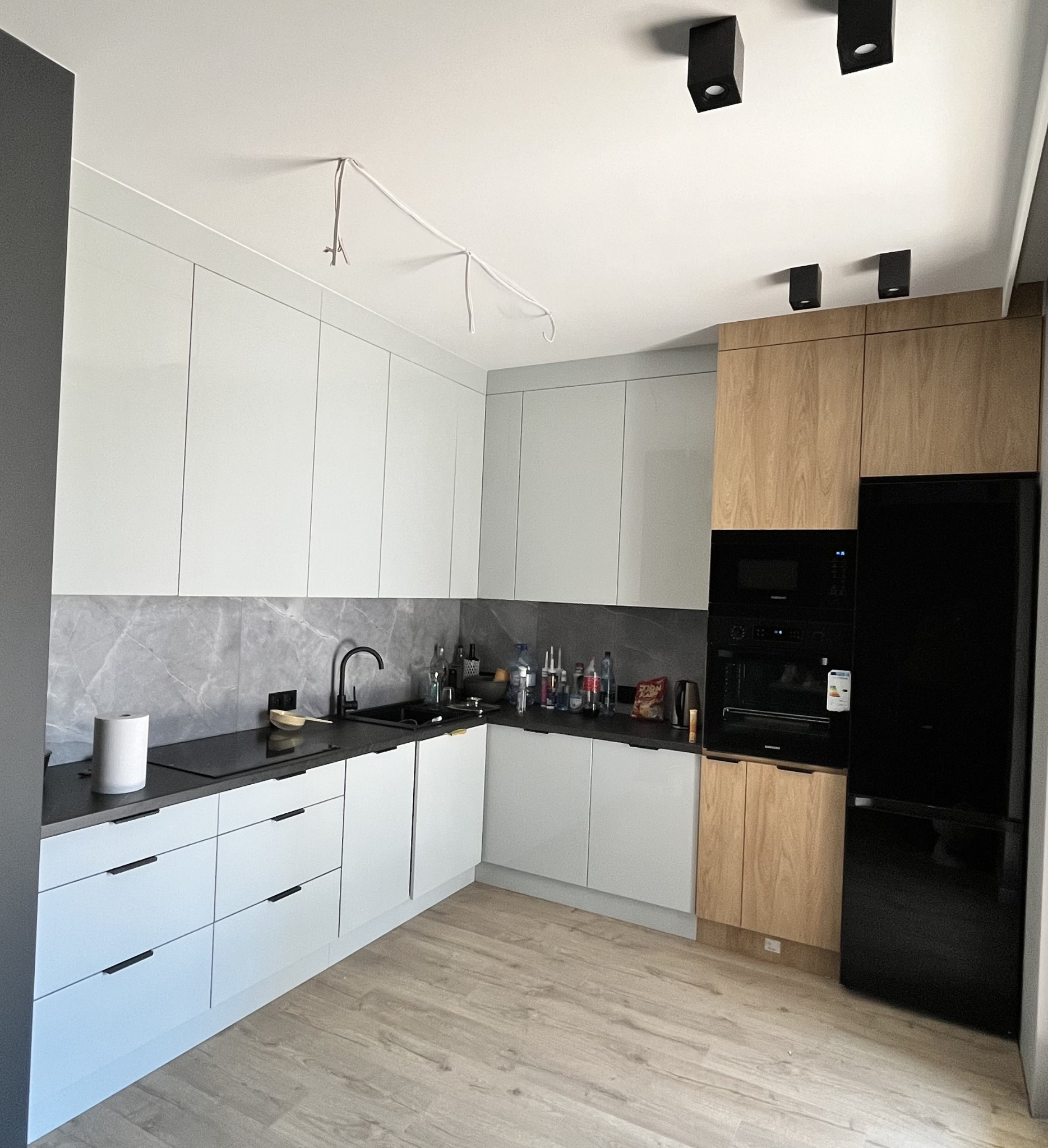 Nowoczesna kuchnia w odcieniach bieli i drewna z czarnymi akcentami, widoczne niedokończone podłączenie oświetlenia sufitowego