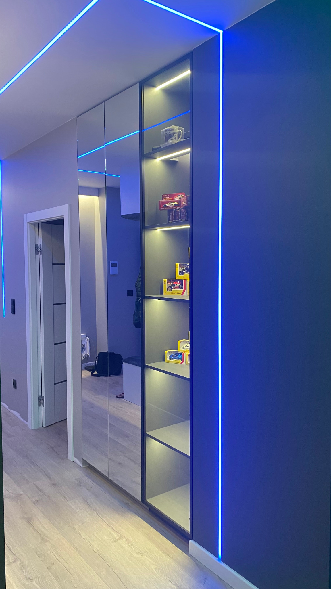 Nowoczesna szafa wnękowa z lustrzanymi drzwiami i podświetleniem LED, eksponująca kolekcję modeli samochodów na półkach.