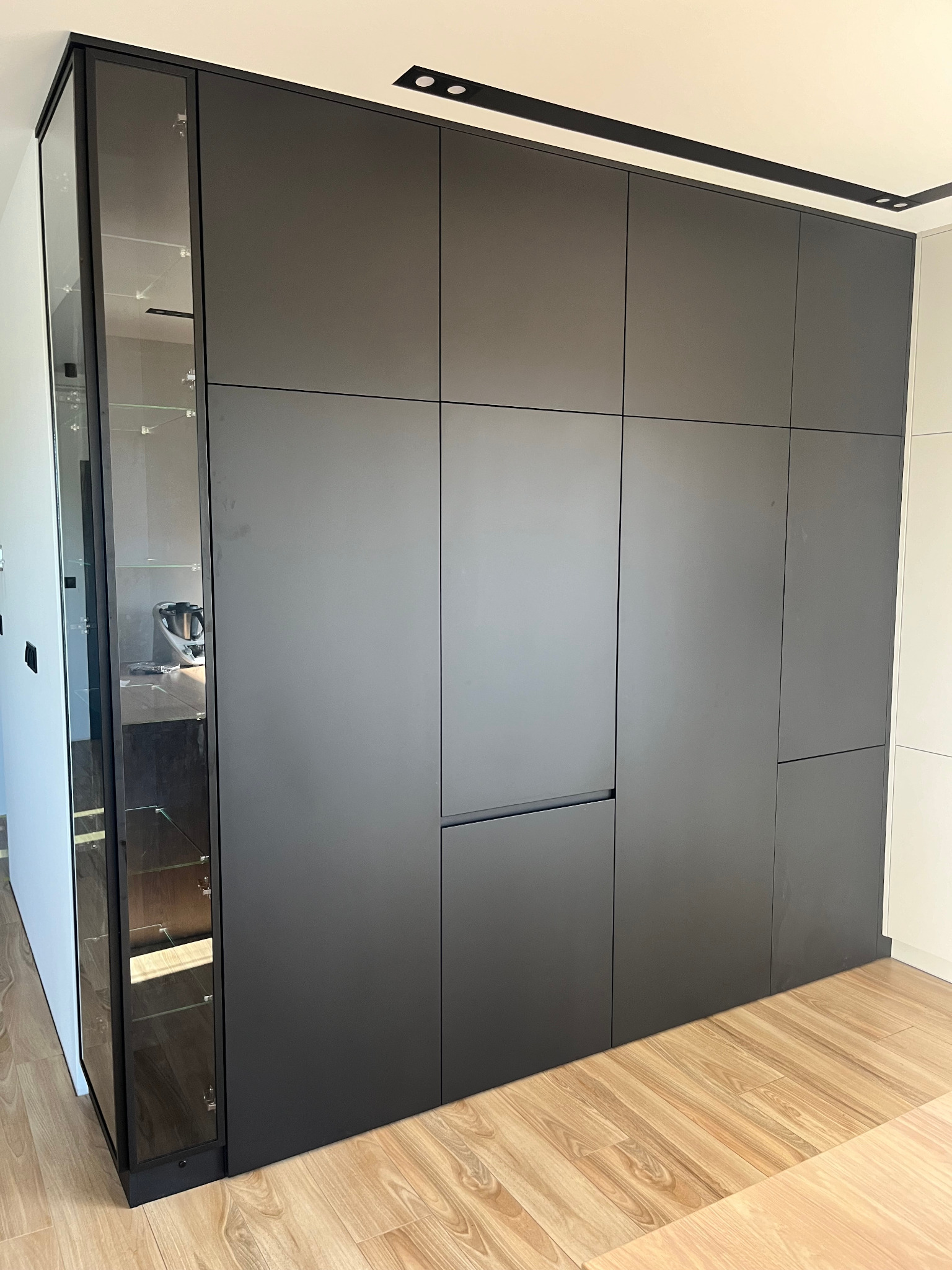 Nowoczesna, matowa szafa w zabudowie kuchennej w odcieniach szarości, z przeszklonym segmentem bocznym i drewnianą podłogą. Minimalistyczny design.