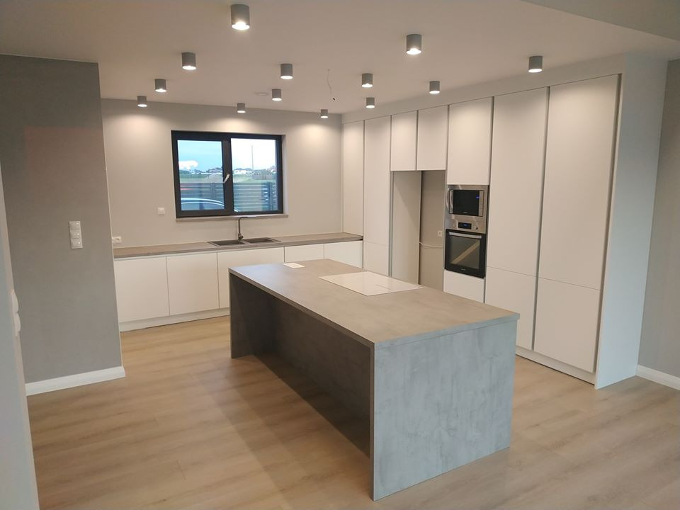 Nowoczesna kuchnia z wyspą: białe szafki, szary blat, płyta indukcyjna. Minimalistyczny design, oświetlenie punktowe, okno z widokiem na zewnątrz. Betonowa wyspa kuchenna.
