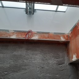 Firma Remontowo - Budowlana PLATRES Rafał Goc - Ściana z nałożonym tynkiem z widocznymi listwami i instalacją elektryczną, przygotowana do dalszych prac wykończeniowych. Widoczne okno dachowe.