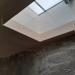 Firma Remontowo - Budowlana PLATRES Rafał Goc - Świeżo położone, szare tynki na ścianie z widocznym oknem dachowym. Widoczne ślady po poprzednich warstwach i wystające przewody elektryczne.