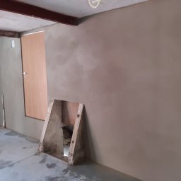 Firma Remontowo - Budowlana PLATRES Rafał Goc - Wnętrze po tynkowaniu maszynowym: gładkie, jasne ściany, widoczny otwór w ścianie z drewnianą obudową, surowy beton na podłodze, metalowa belka pod sufitem.