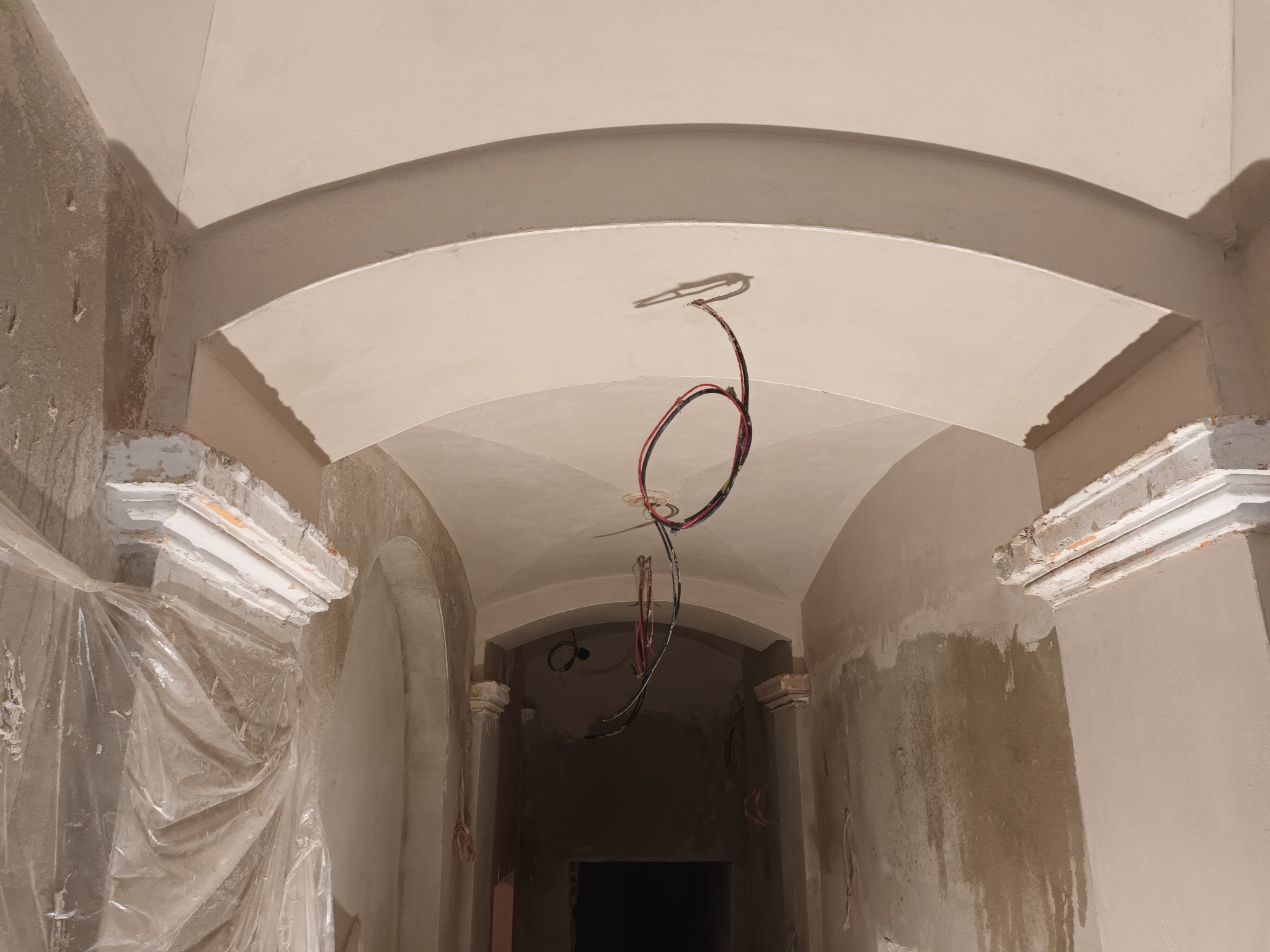 Korytarz w trakcie prac tynkarskich z charakterystycznymi łukami i widocznymi przewodami elektrycznymi. Ściany w odcieniach beżu, częściowo zabezpieczone folią.