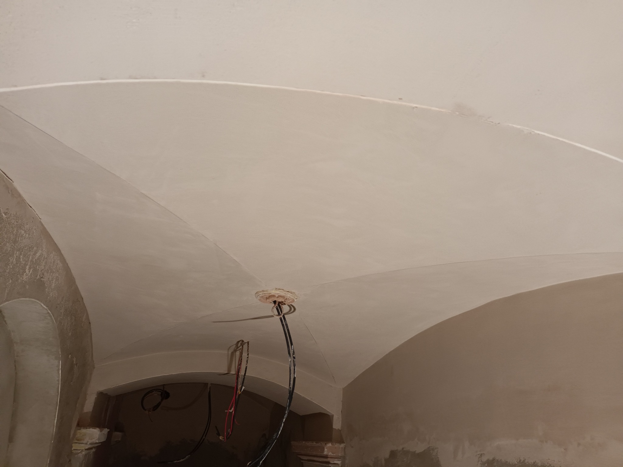 Gładkie, jasne tynki na sklepieniu łukowym z widoczną instalacją elektryczną. Świeżo położone tynki w odcieniach beżu i bieli. Widoczne fragmenty ścian i łuków.