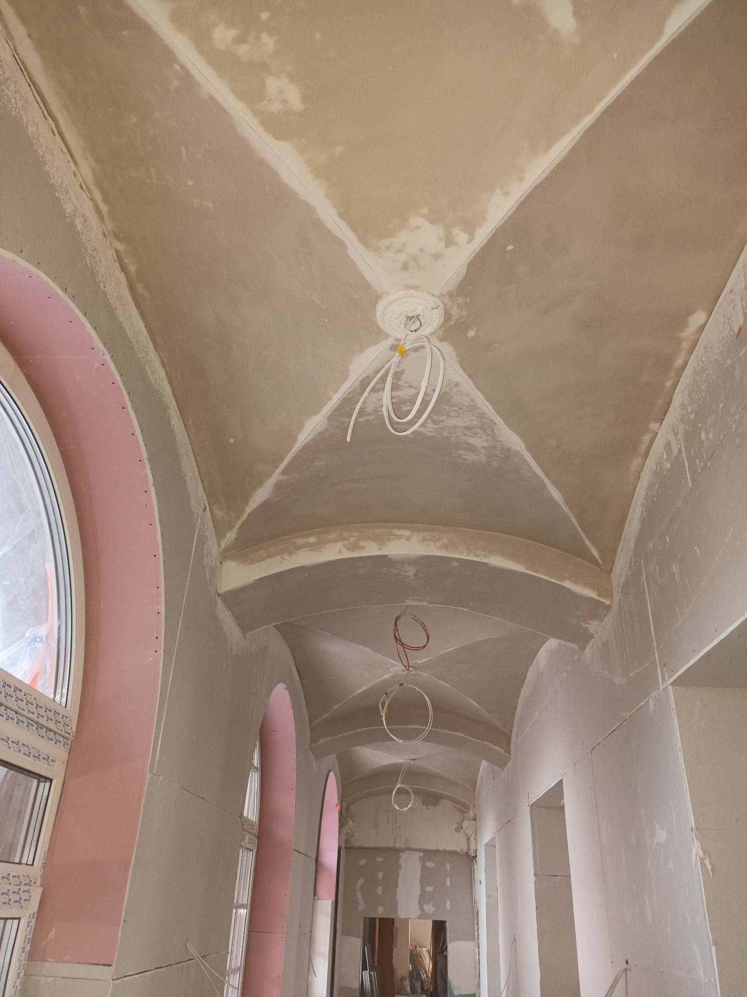 Korytarz w trakcie prac wykończeniowych: sufit z łukami, widoczne przewody elektryczne, ściany przygotowane pod tynk. Okna z różowymi ramami. Wnętrze w stanie surowym.