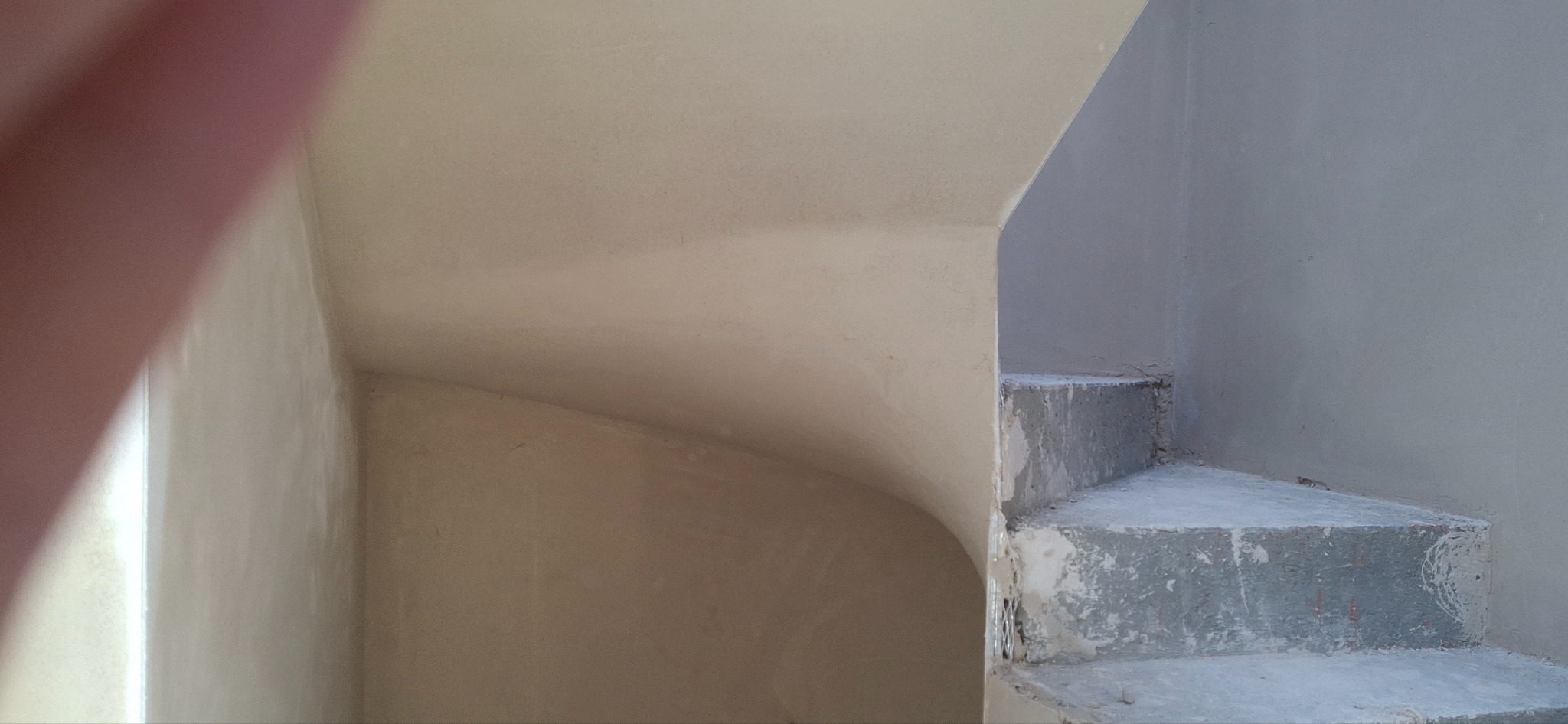 Wnętrze po tynkowaniu maszynowym: gładkie ściany i schody w surowym stanie, gotowe do dalszych prac wykończeniowych. Widoczne zaokrąglenie ściany.