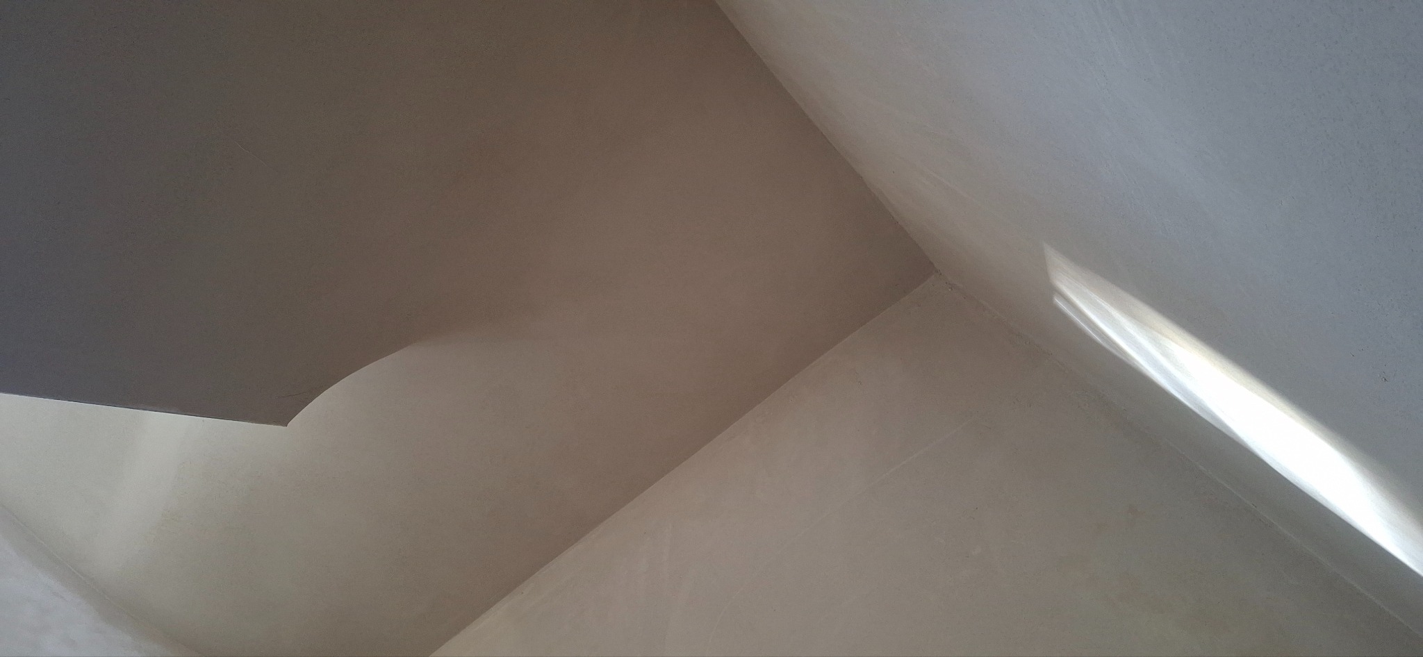 Gładka, jasna powierzchnia ściany po tynkowaniu maszynowym, z widocznym załamaniem światła w rogu pomieszczenia i fragmentem sufitu. Minimalistyczne ujęcie.