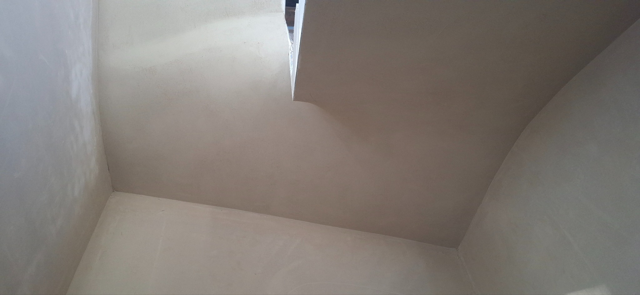 Gładkie, jasne tynki na ścianach i suficie w rogu pomieszczenia. Widoczne delikatne zaokrąglenia sufitu. Minimalistyczne wykończenie wnętrza.