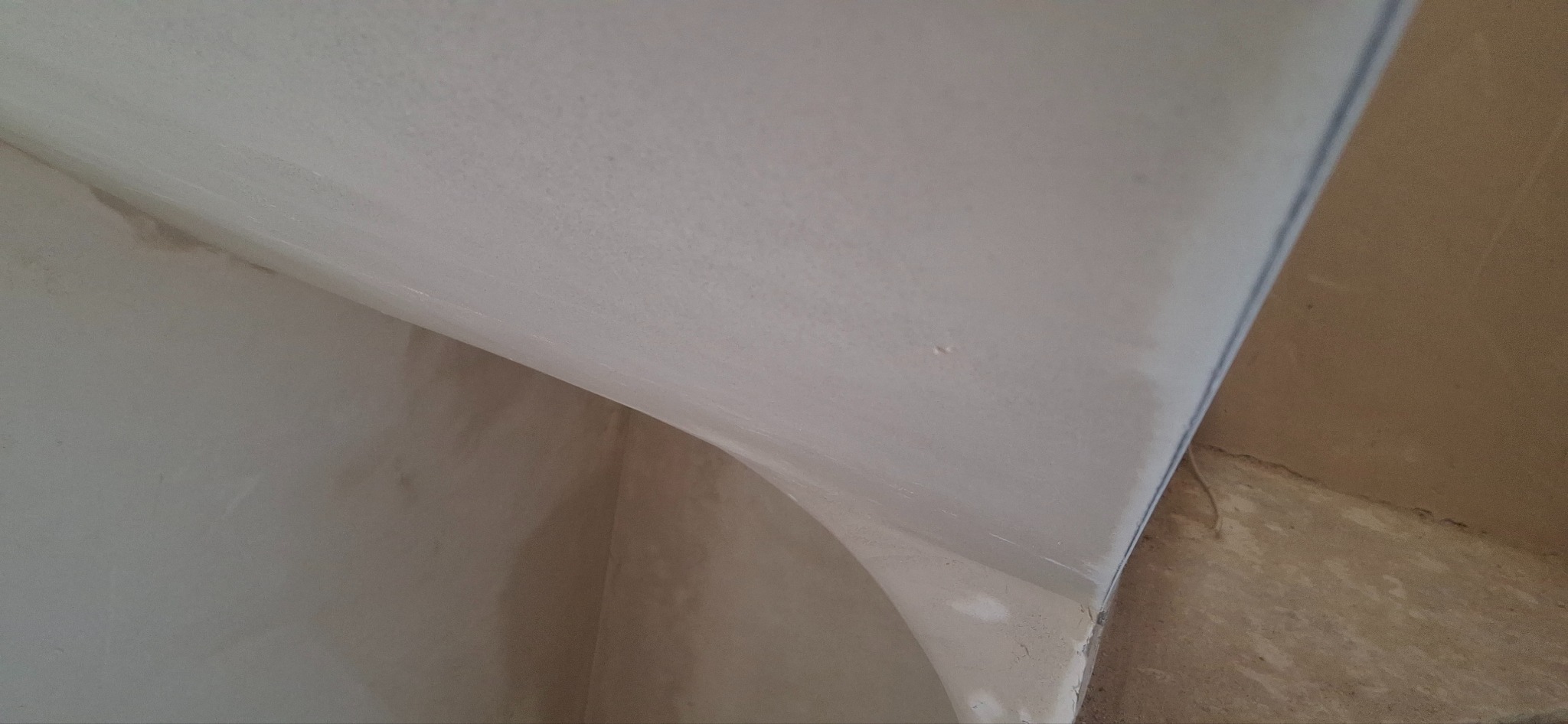 Gładka, jasna powierzchnia ściany po tynkowaniu maszynowym, z widocznym zaokrągleniem narożnika i delikatną fakturą. Ujęcie z dołu, podkreślające precyzję wykonania.