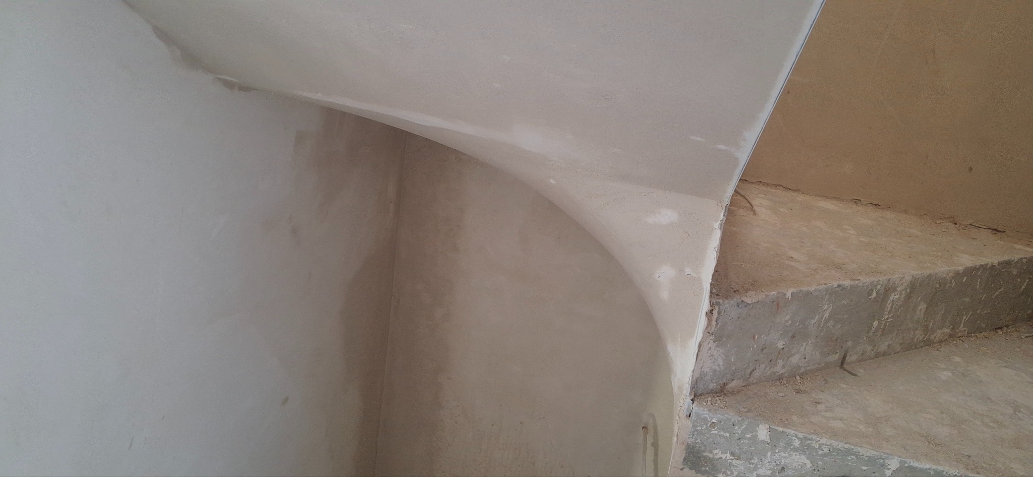 Ściana po tynkowaniu maszynowym z zaokrąglonym narożnikiem i fragmentem schodów w surowym stanie. Widoczne niedoskonałości i pozostałości po pracach budowlanych.