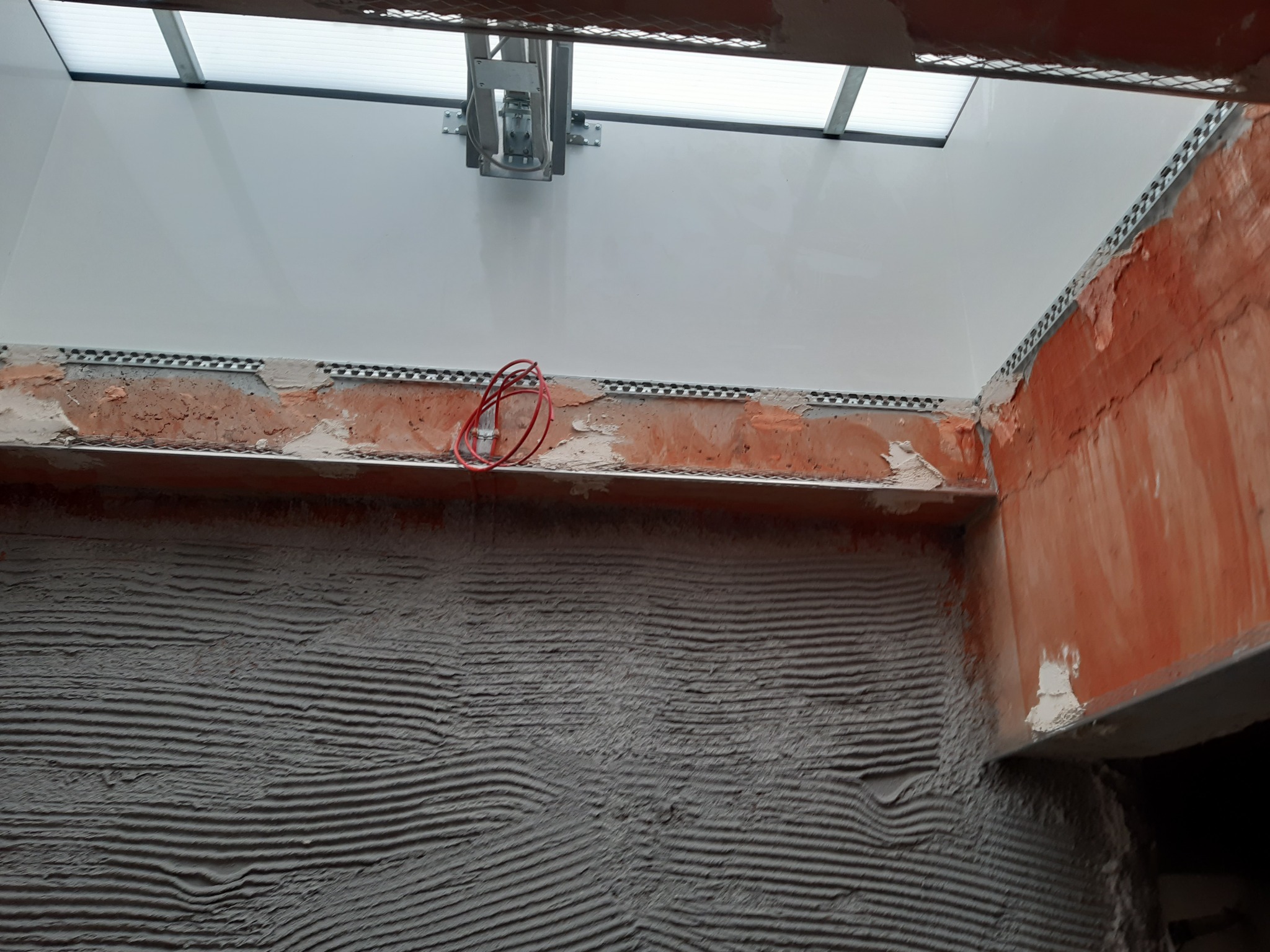 Ściana z nałożonym tynkiem z widocznymi listwami i instalacją elektryczną, przygotowana do dalszych prac wykończeniowych. Widoczne okno dachowe.