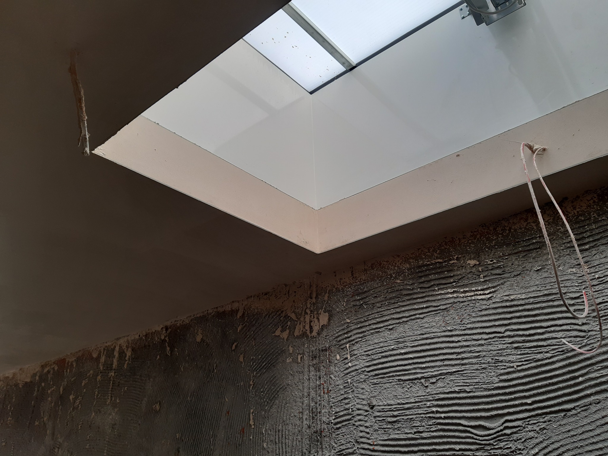 Świeżo położone, szare tynki na ścianie z widocznym oknem dachowym. Widoczne ślady po poprzednich warstwach i wystające przewody elektryczne.