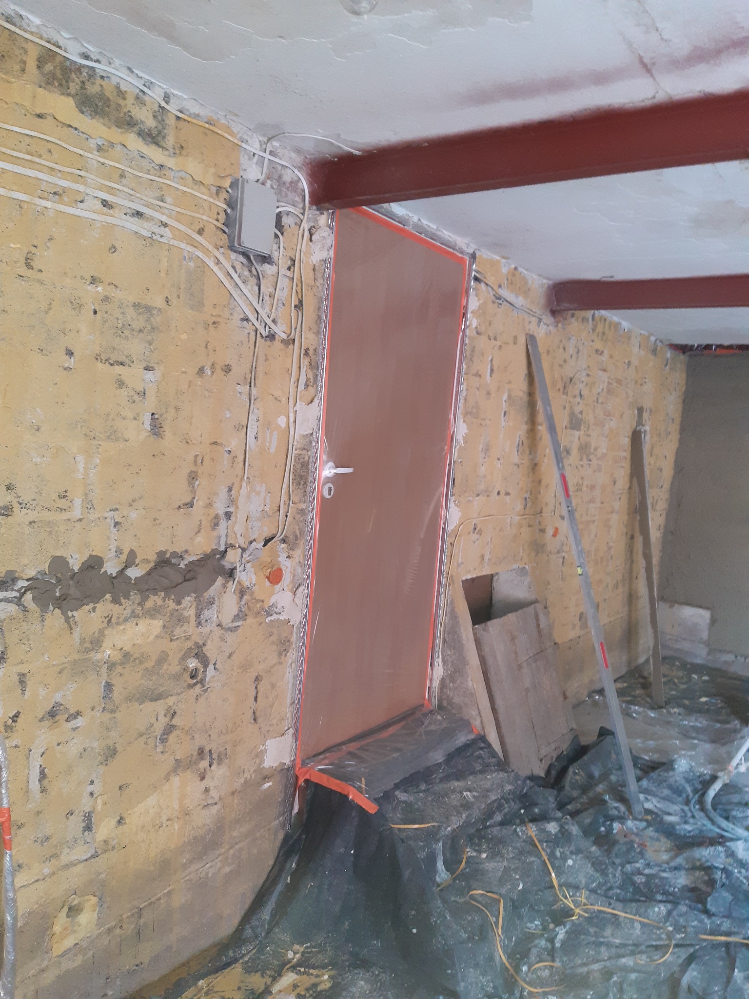 Przygotowanie do tynkowania: surowa ściana z widocznymi instalacjami elektrycznymi, zabezpieczona folią podłoga i osłonięte drzwi. Widoczne poziomice i stalowa belka stropowa.