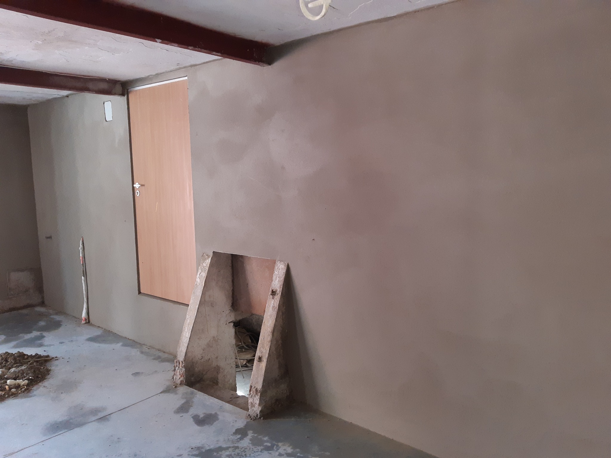 Wnętrze po tynkowaniu maszynowym: gładkie, jasne ściany, widoczny otwór w ścianie z drewnianą obudową, surowy beton na podłodze, metalowa belka pod sufitem.