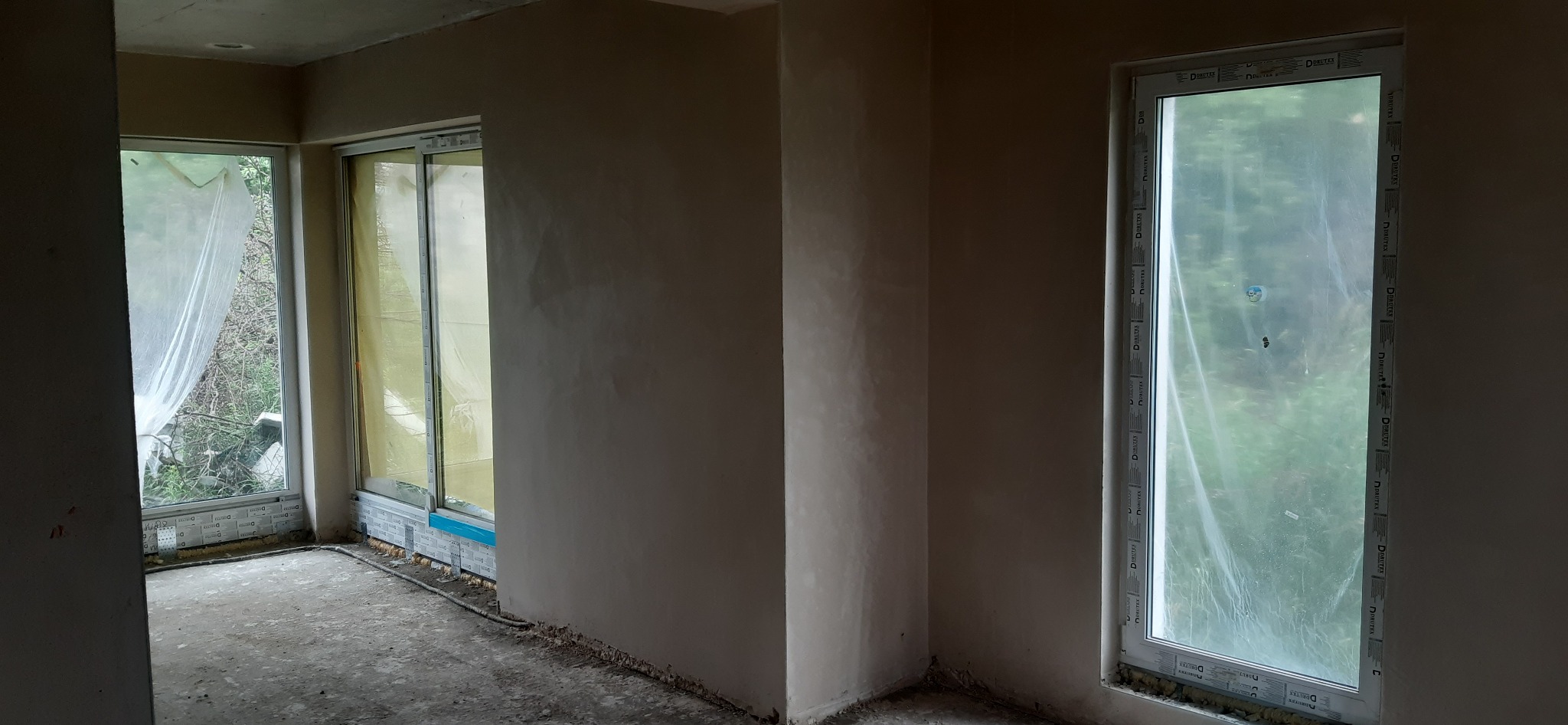 Wnętrze po tynkowaniu: gładkie, jasne ściany, okna zabezpieczone folią. Widoczne ślady prac wykończeniowych na podłodze. Minimalistyczny kadr, surowy stan.
