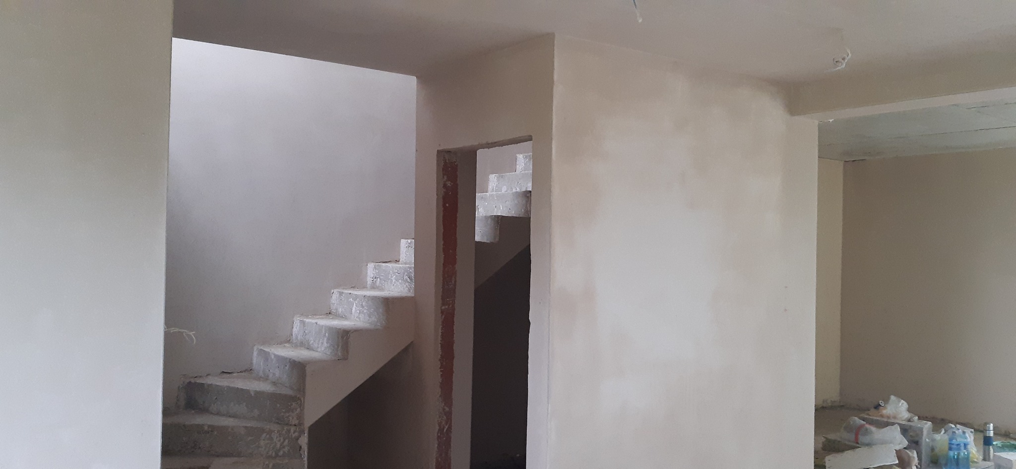 Wnętrze po tynkowaniu: gładkie, jasne ściany, surowe schody betonowe. Widoczne ślady prac wykończeniowych i materiały budowlane. Minimalistyczny kadr.