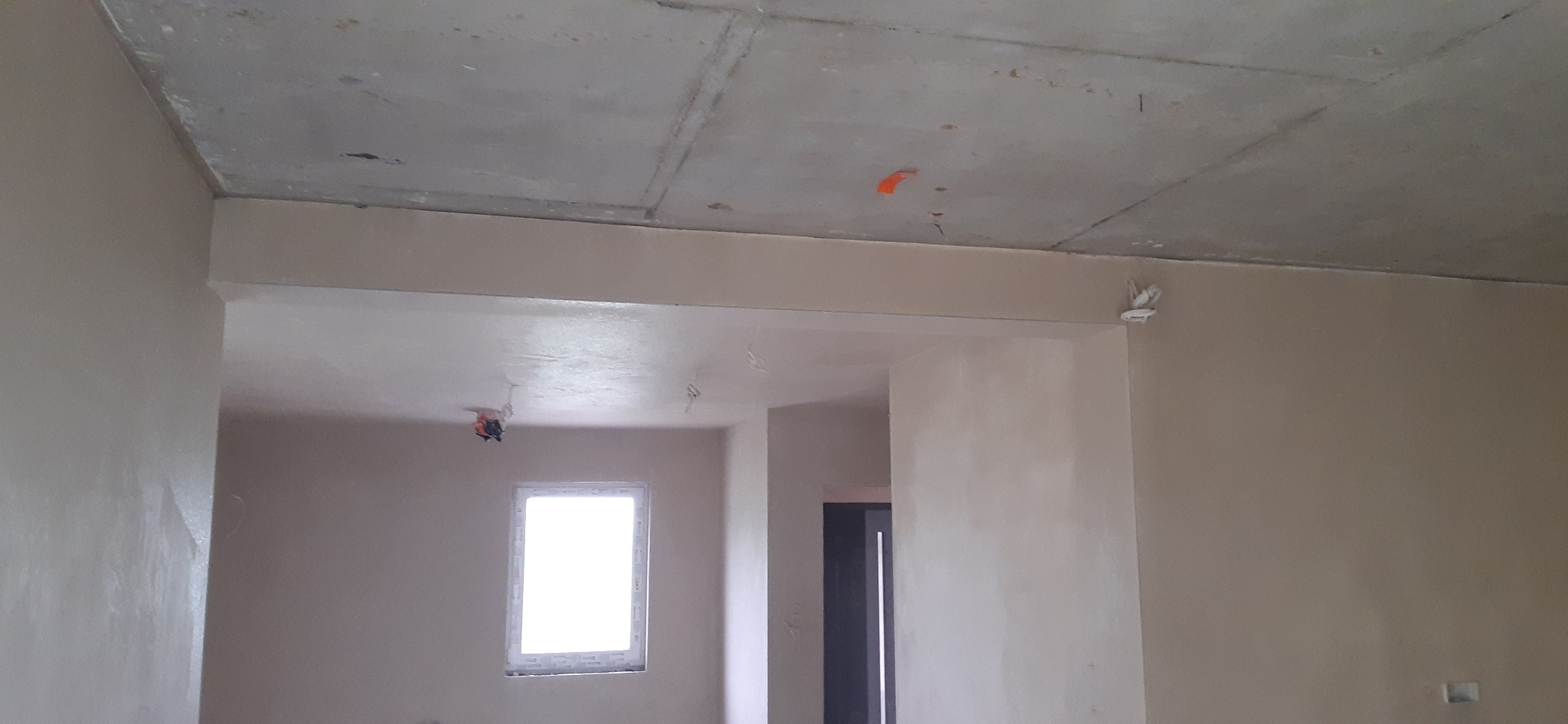 Wnętrze po tynkowaniu maszynowym: gładkie ściany w jasnym odcieniu, surowy betonowy sufit z widocznymi instalacjami. Widok na okno i przejście do innego pomieszczenia.