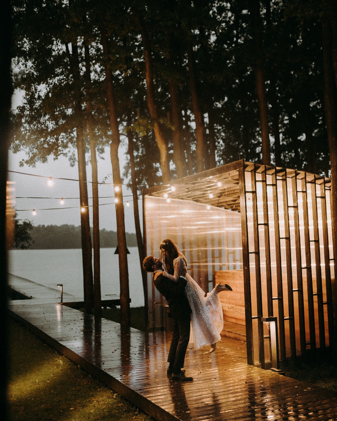 Para młoda w objęciach na mokrym drewnianym pomoście oświetlonym girlandami żarówek, jezioro w tle, ciemne drzewa i konstrukcja z przezroczystymi ścianami.