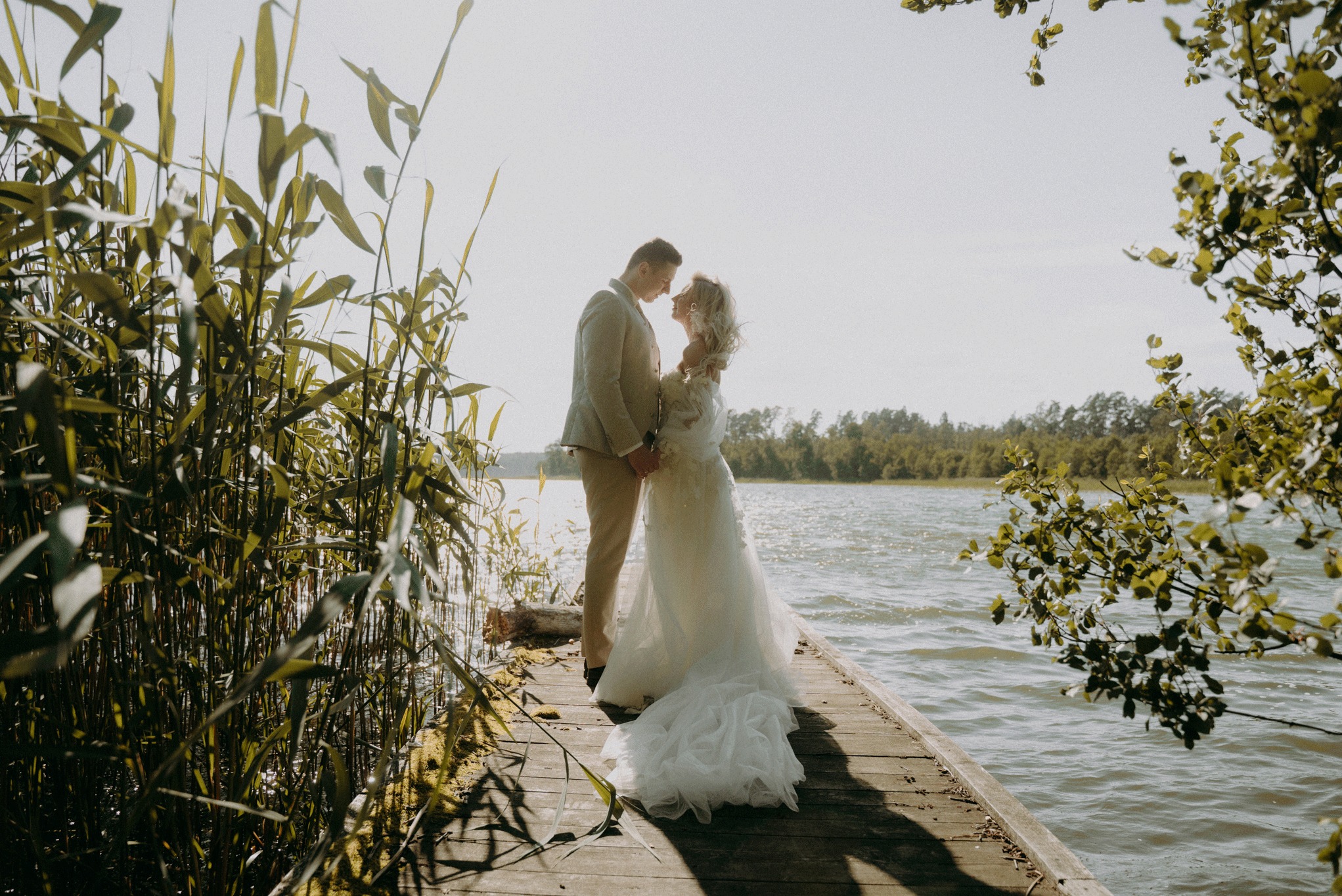 Para młoda na drewnianym pomoście nad jeziorem, panna młoda w koronkowej sukni z trenem, pan młody w beżowym garniturze, otoczeni trzcinami i zielenią.