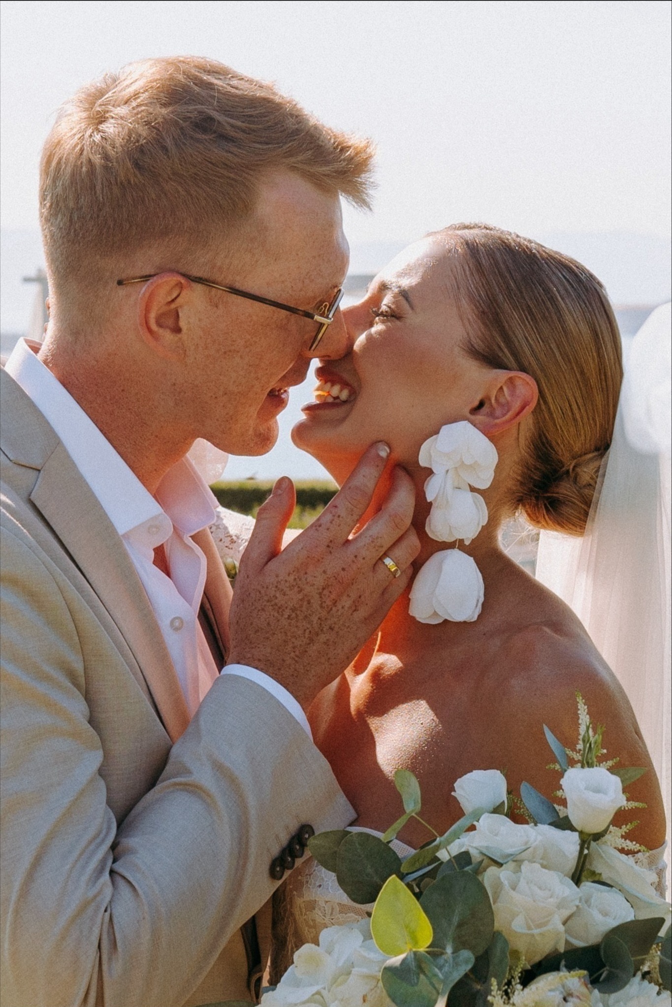 Uśmiechnięta para młoda w beżowych strojach, zbliżona do pocałunku, panna młoda w dużych, białych kolczykach kwiatowych i z bukietem białych róż.