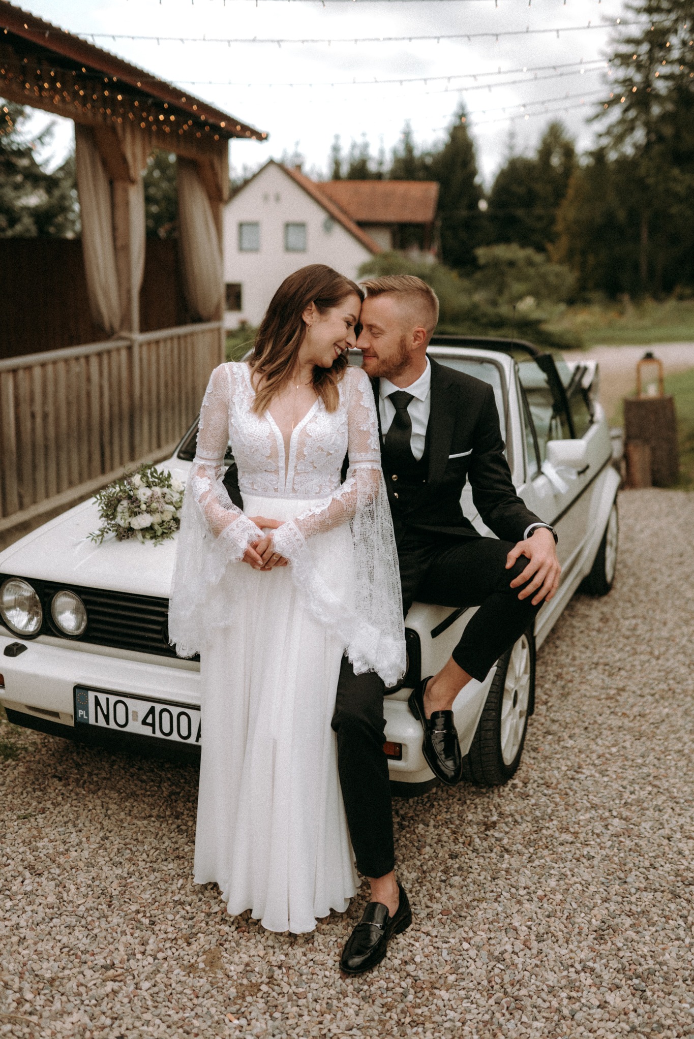 Para młoda, panna młoda w koronkowej sukni z bukietem, pan młody w czarnym garniturze, siedząca na białym kabriolecie z numerem rejestracyjnym NO 400A, uśmiechnięci, dotykający się czołami, w tle...