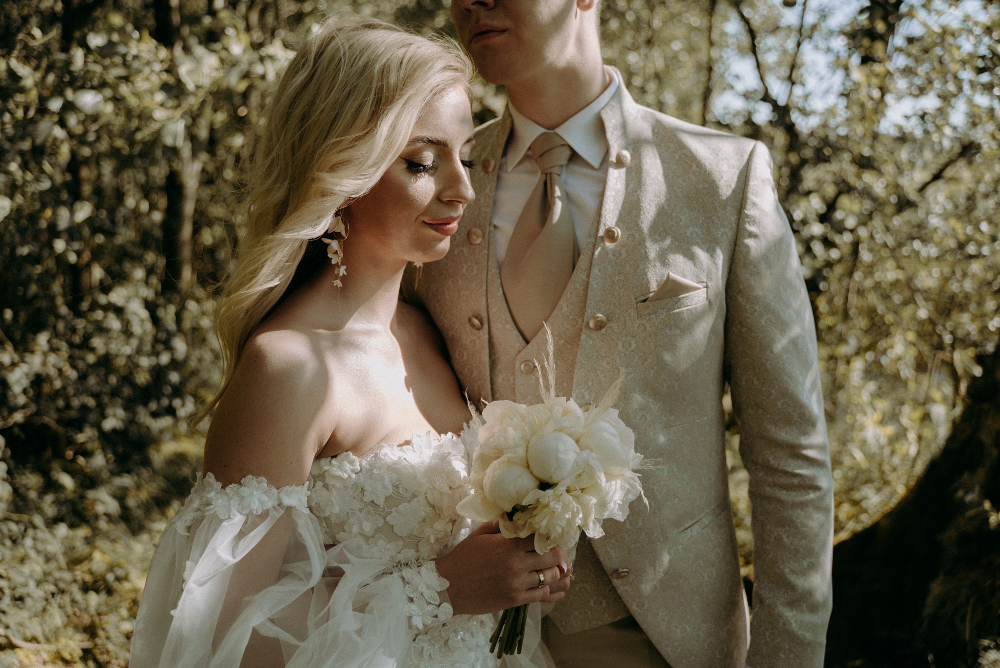 Panna młoda w sukni ślubnej z kwiatowymi zdobieniami i bukietem białych piwonii, obok pan młody w beżowym garniturze z wzorem paisley, na tle zieleni drzew.