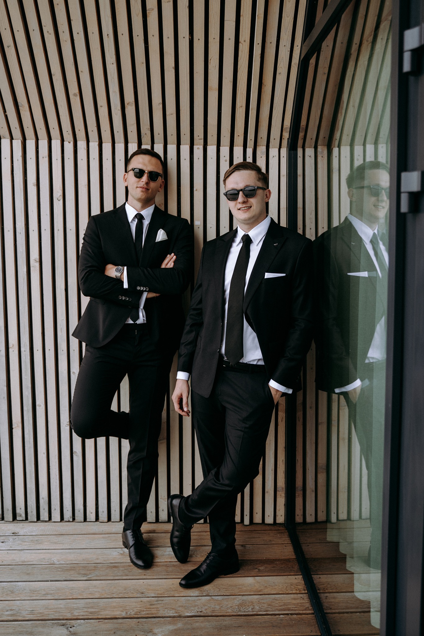 Dwóch elegancko ubranych mężczyzn w czarnych garniturach i okularach przeciwsłonecznych pozuje na tle drewnianej ściany i szklanej witryny, jeden ze skrzyżowanymi ramionami, drugi z ręką w kieszeni.