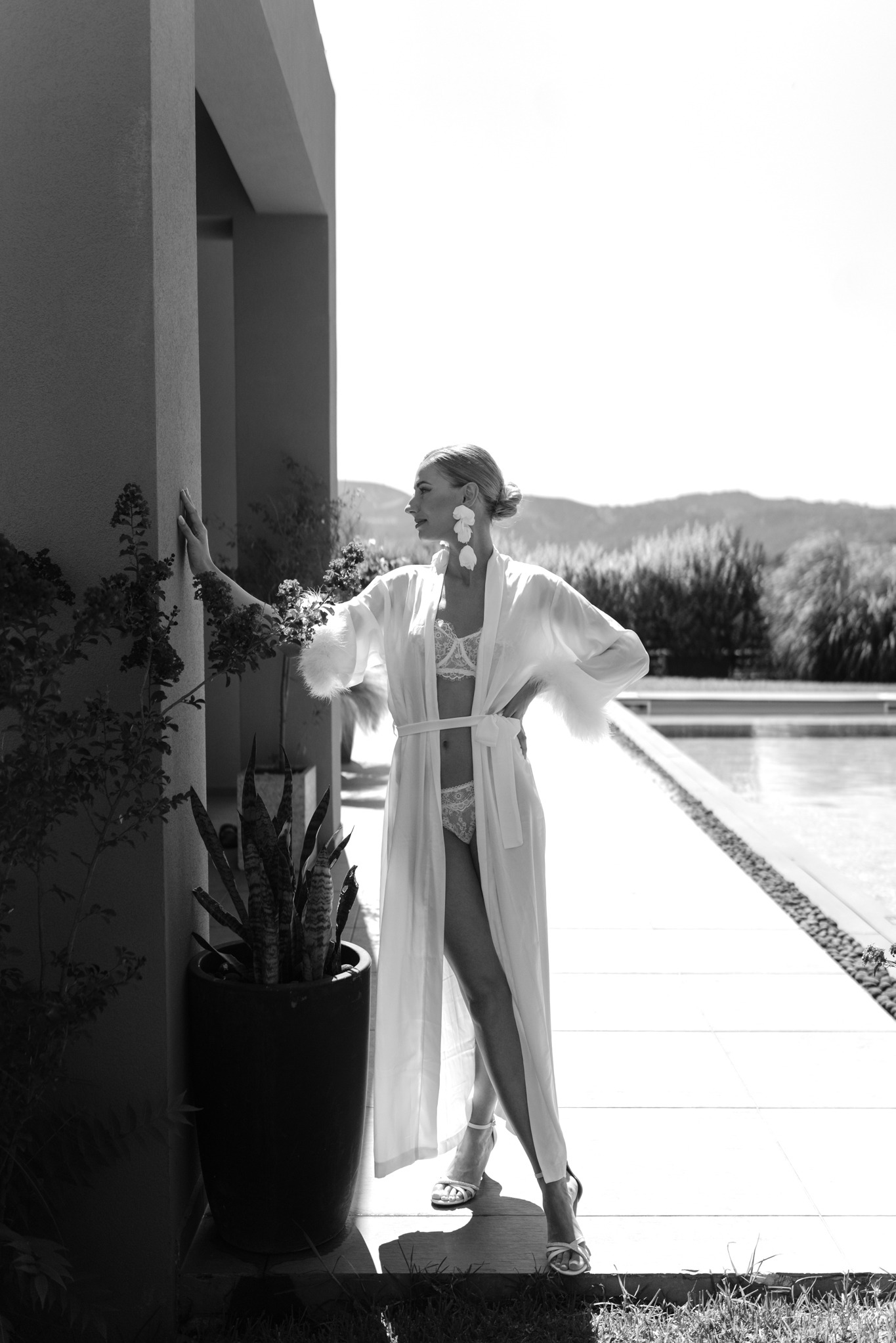 Kobieta w bieliźnie i szlafroku z piórami, pozująca przy basenie w słoneczny dzień. Ujęcie w czerni i bieli.