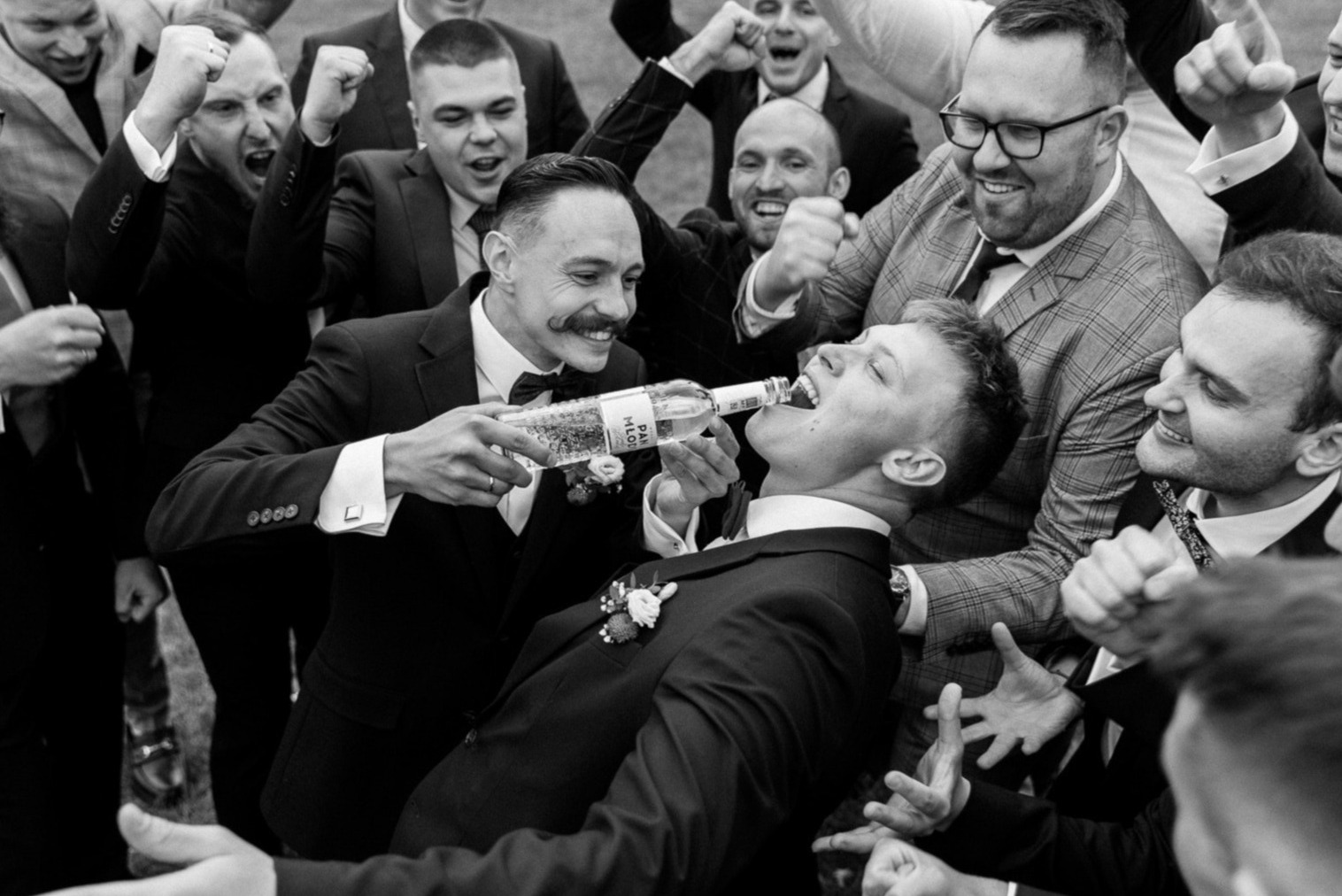 Czarno-białe zdjęcie z wesela: mężczyzna z wąsami nalewa wódkę do ust leżącego pana młodego, otoczeni przez wiwatujących gości w garniturach. Radosna, spontaniczna chwila.