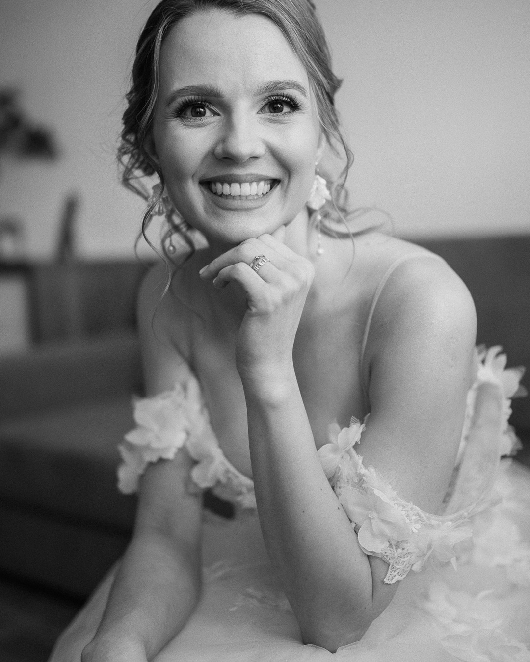 Portret uśmiechniętej panny młodej w sukni z kwiatowymi aplikacjami, opierającej podbródek na dłoni z widocznym pierścionkiem zaręczynowym. Czarno-biała fotografia.