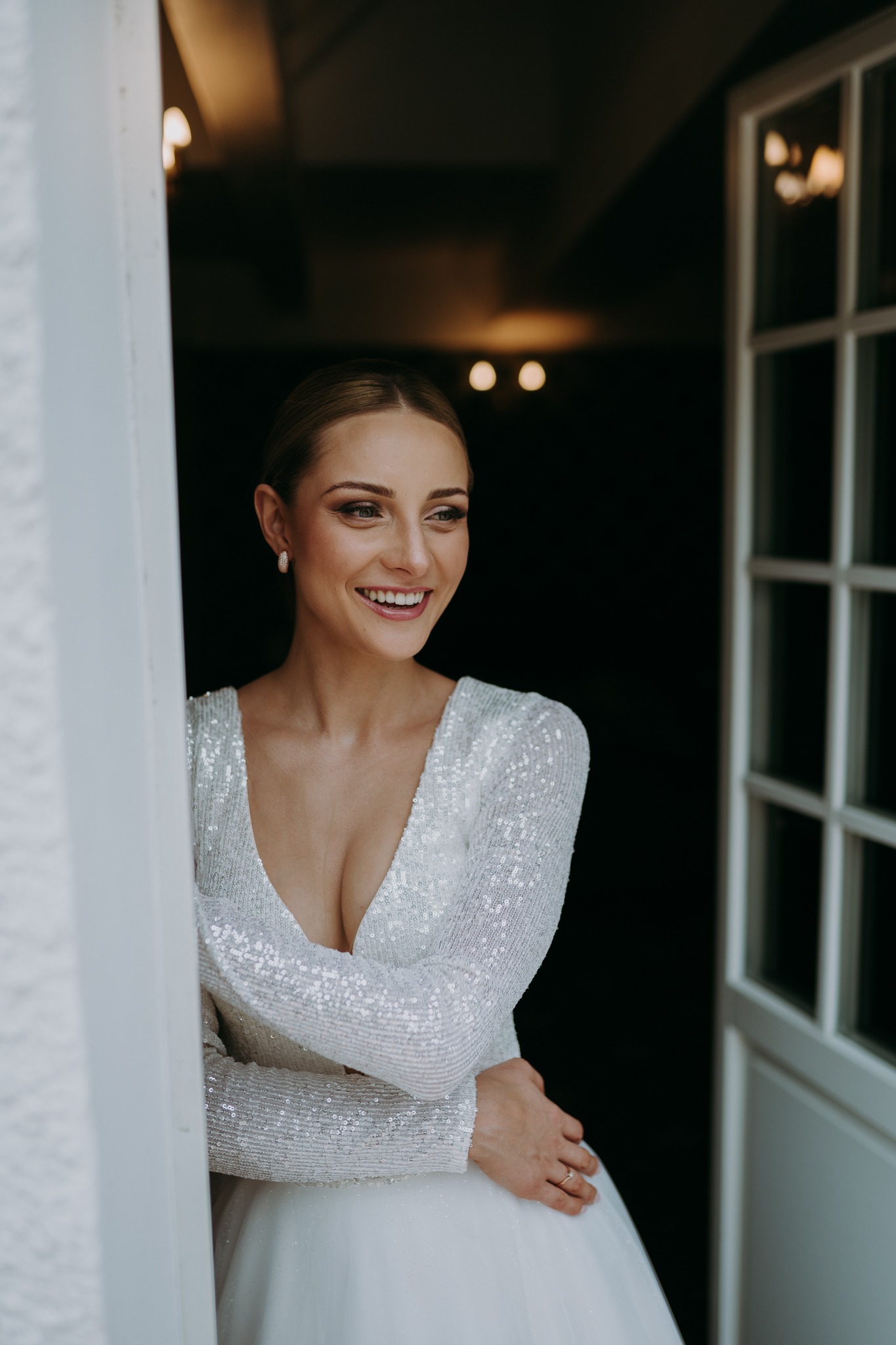 Uśmiechnięta panna młoda w błyszczącej sukni, spogląda w bok, opierając się o białe drzwi. Delikatne oświetlenie wnętrza i naturalny makijaż podkreślają jej urodę.