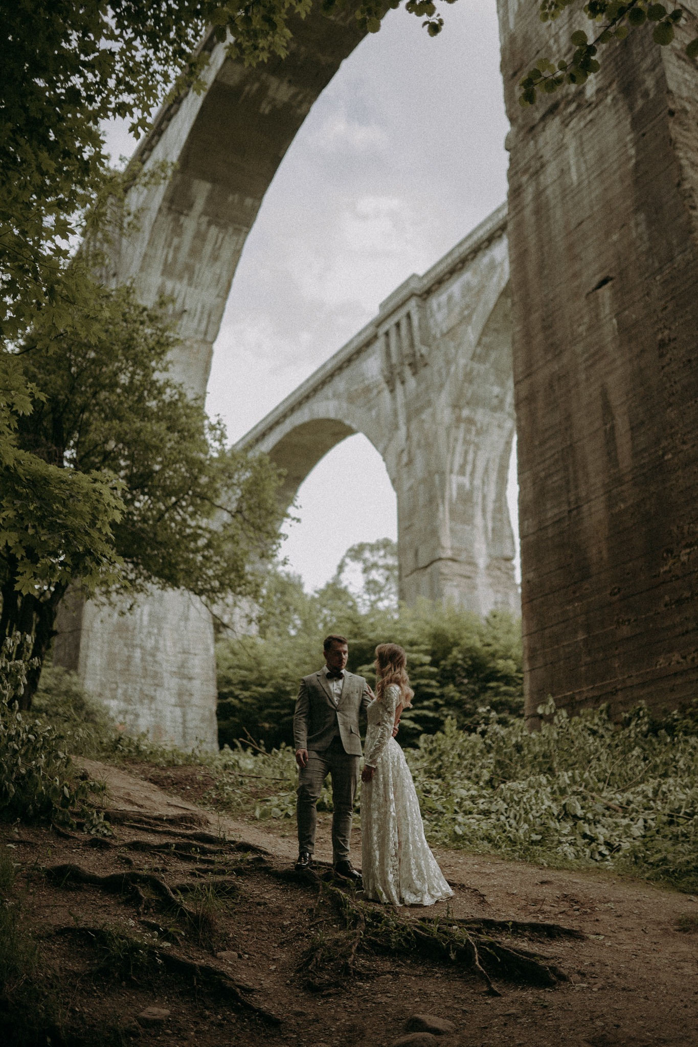 Para młoda w leśnej scenerii pod monumentalnym wiaduktem. Pan w szarym garniturze, pani w koronkowej sukni. Romantyczna sesja w plenerze, unikalne tło.
