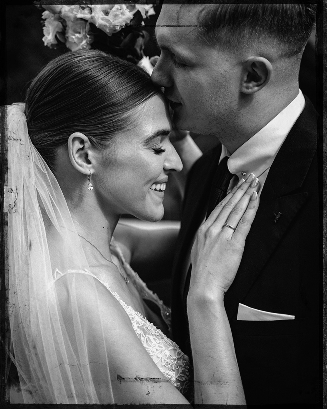 Czarno-białe, portretowe ujęcie młodej pary w dniu ślubu: uśmiechnięta panna młoda z welonem i pan młody w garniturze, czule dotykający czołem jej głowy.