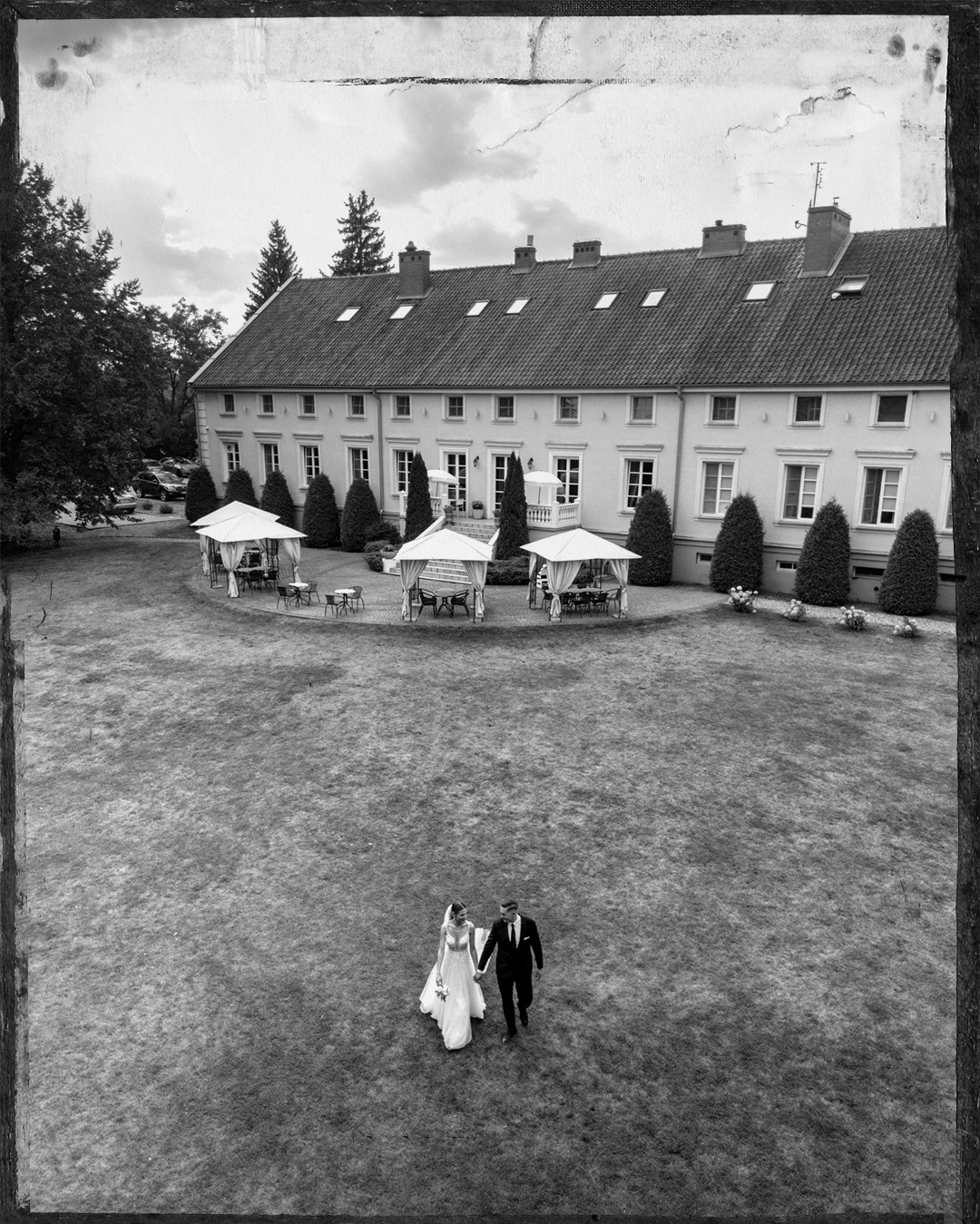 Czarno-białe ujęcie z góry: para młoda spaceruje po trawniku przed budynkiem z licznymi oknami i altanami. Efekt vintage, stylizowane na stare zdjęcie.