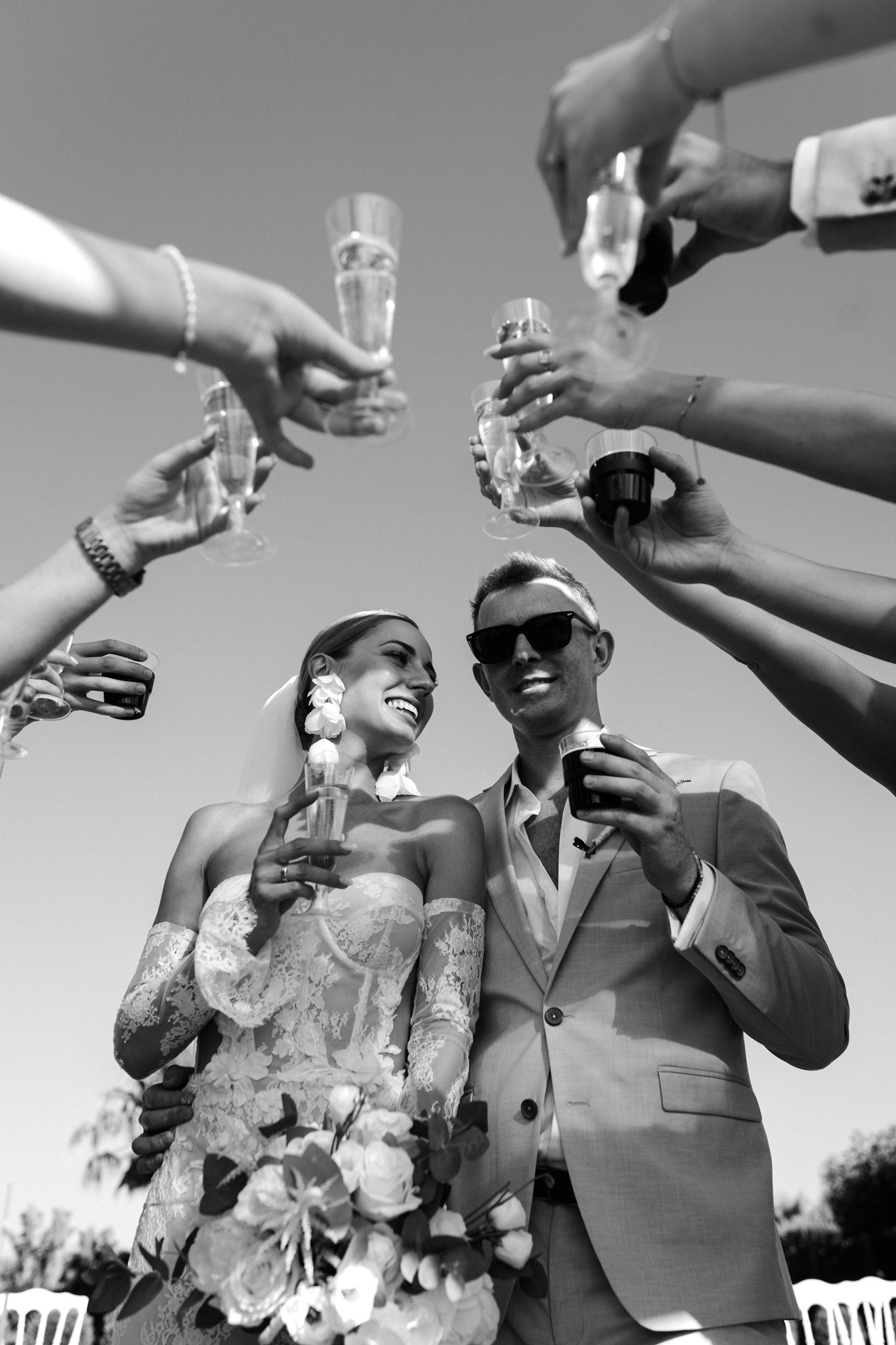 Czarno-białe ujęcie nowożeńców wznoszących toast z gośćmi. Panna młoda w koronkowej sukni, pan młody w garniturze i okularach. Kąt od dołu podkreśla radość.