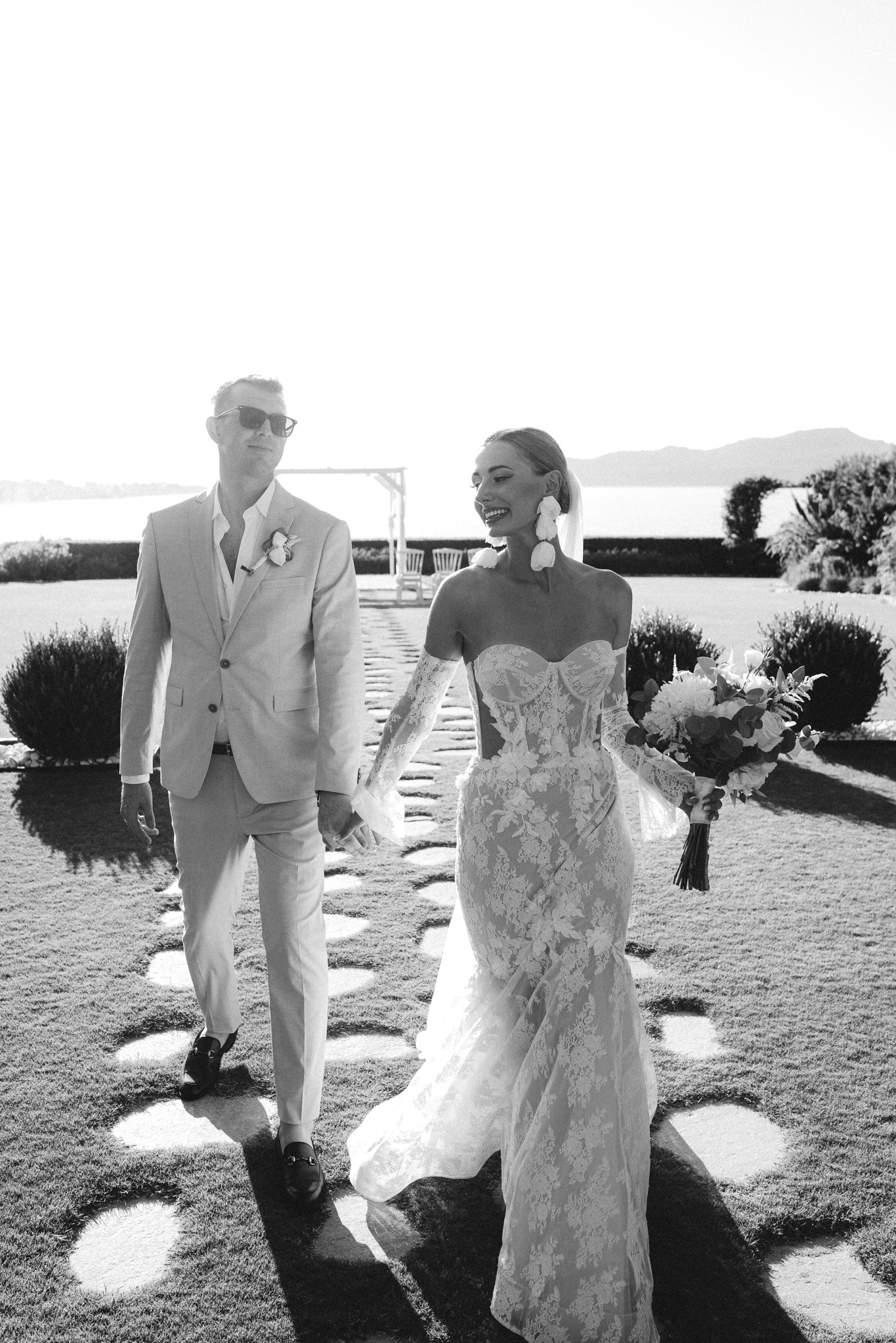 Czarno-białe zdjęcie pary młodej idącej po trawie. Panna młoda w koronkowej sukni, pan młody w jasnym garniturze i okularach. W tle morze i góry.