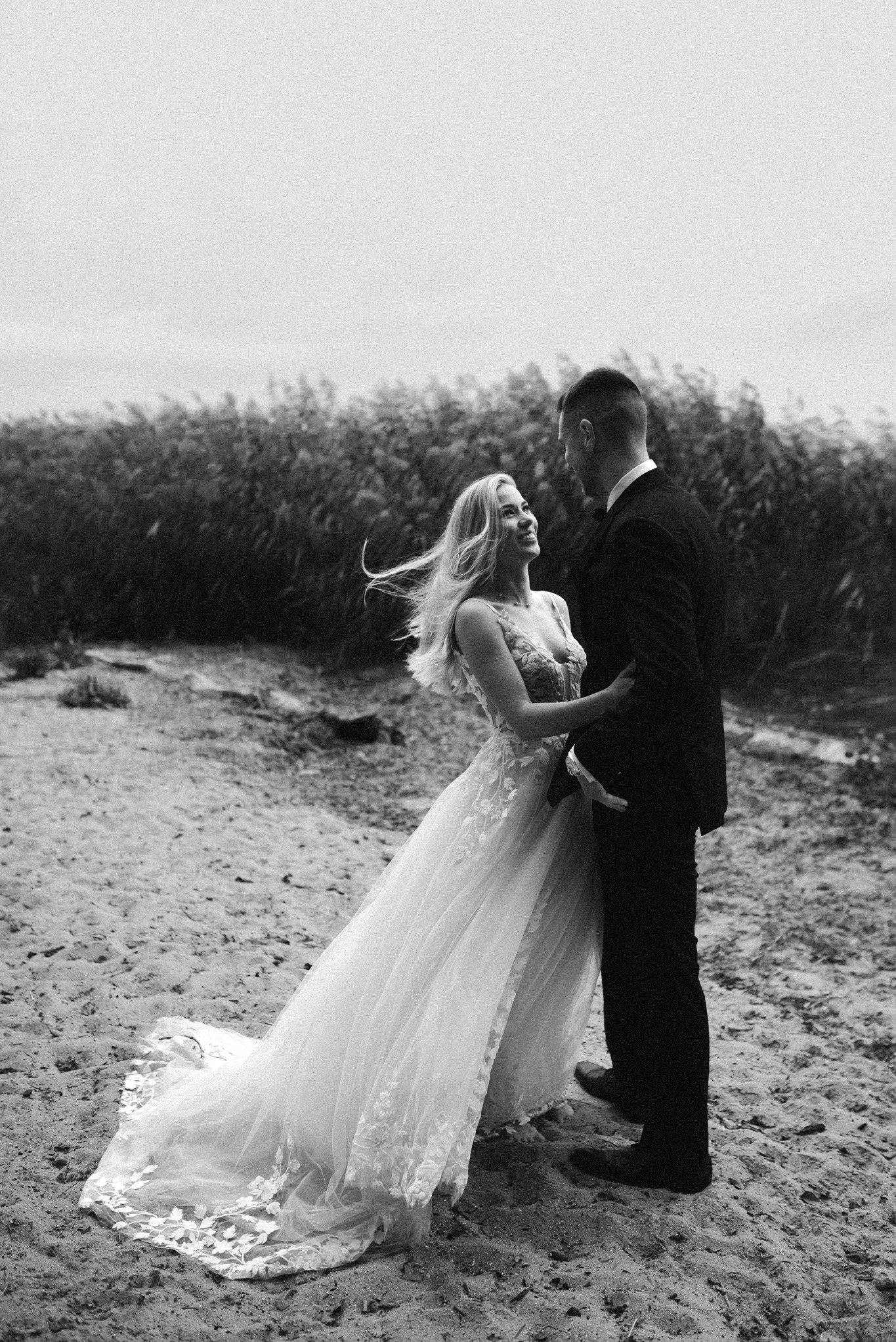 Czarno-białe zdjęcie pary młodej na plaży: panna młoda w koronkowej sukni patrzy z uśmiechem na pana młodego w garniturze, w tle szuwary. Wietrzna pogoda.