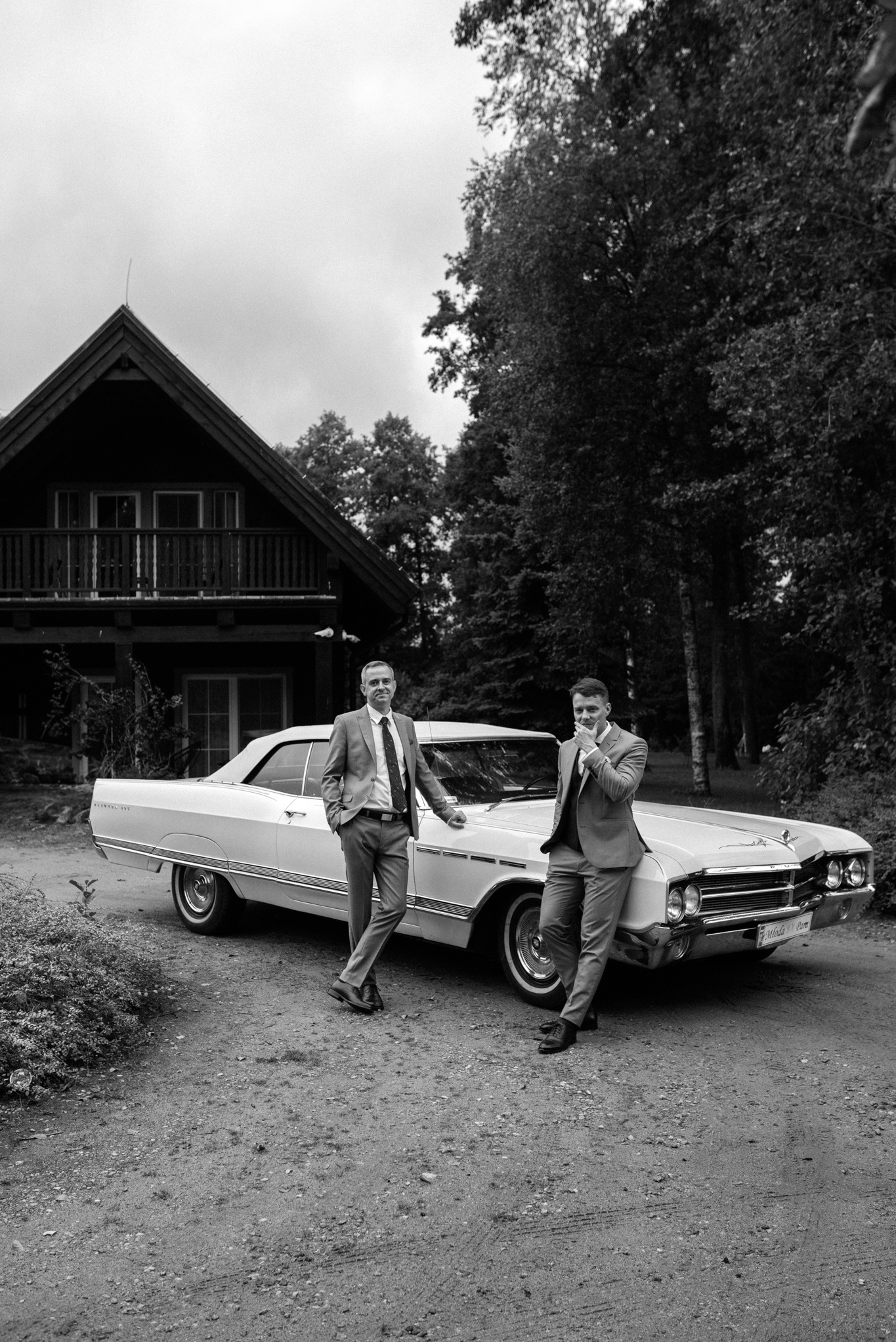 Czarno-białe zdjęcie dwóch eleganckich mężczyzn w garniturach opartych o biały, zabytkowy kabriolet na tle drewnianego domu i drzew. Tablica rejestracyjna z napisem 'Młoda Para'.