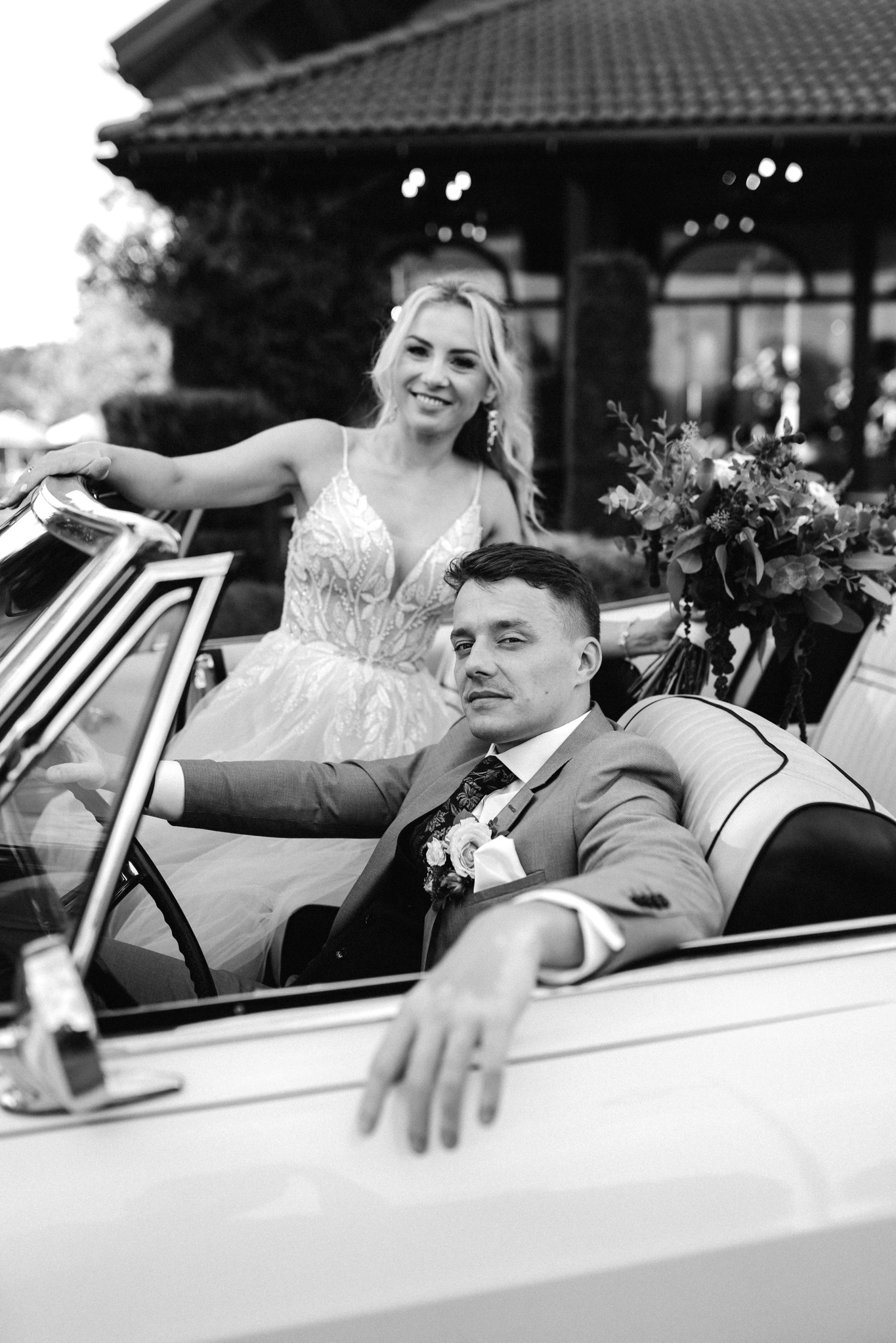 Czarno-białe zdjęcie pary młodej w kabriolecie w Olecku. Pan młody za kierownicą, panna młoda opiera się o auto, oboje uśmiechnięci, w tle budynek.