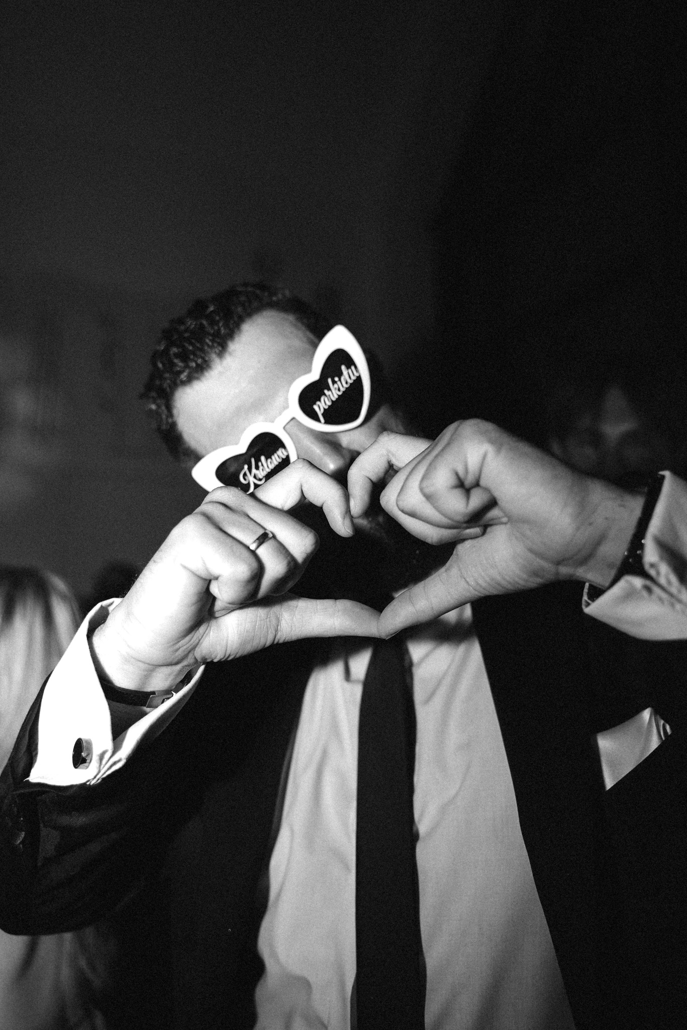 Czarno-białe zdjęcie mężczyzny w okularach w kształcie serc z napisem 'Kidawa' i 'Parkietu', dłońmi formującego serce. Elegancki strój, mankiety koszuli.