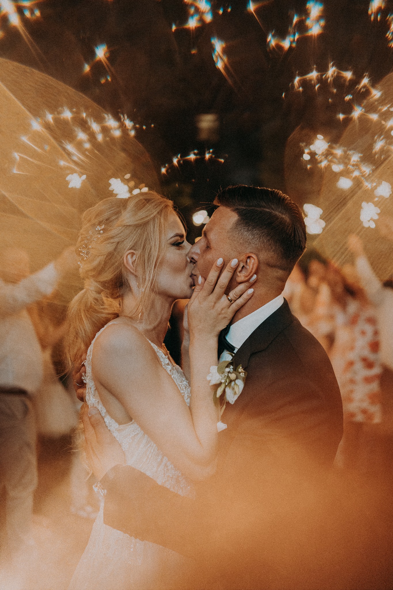 Pocałunek pary młodej w otoczeniu świateł podczas wesela. Panna młoda w sukni z koronką, pan młody w ciemnym garniturze z butonierką. Ciepłe światło i bokeh.