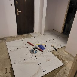 Mateusz Trojan - Układanie jasnych, marmurkowych płytek podłogowych w korytarzu, widoczne narzędzia glazurnicze i poziomica, fuga jeszcze nie położona.