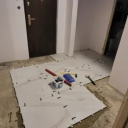 Układanie jasnych, marmurkowych płytek podłogowych w korytarzu, widoczne narzędzia glazurnicze i poziomica, fuga jeszcze nie położona.