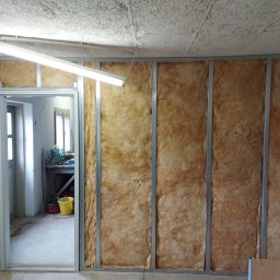 Mateusz Trojan - Ściana w trakcie izolacji wełną mineralną, widoczne profile metalowe i fragment otwartych drzwi, w tle żółte wiadro