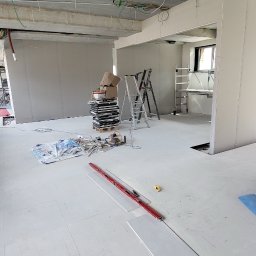 Mateusz Trojan - Wnętrze w trakcie remontu: widoczne płyty gipsowo-kartonowe na ścianach, surowy betonowy sufit z instalacjami, ułożone panele podłogowe, drabiny i narzędzia.