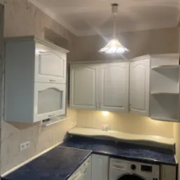 Jasna kuchnia z białymi szafkami, niebieskim blatem i wzorzystą tapetą, oświetlona wiszącą lampą i podświetleniem pod szafkami.