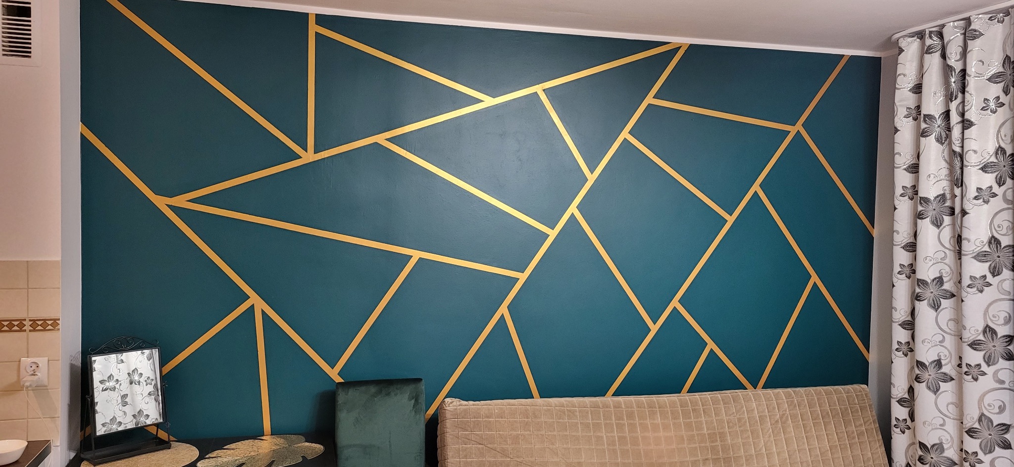 Turkusowa ściana z geometrycznym wzorem z cienkich, złotych linii, widoczny fragment beżowej sofy i zasłony w kwiaty